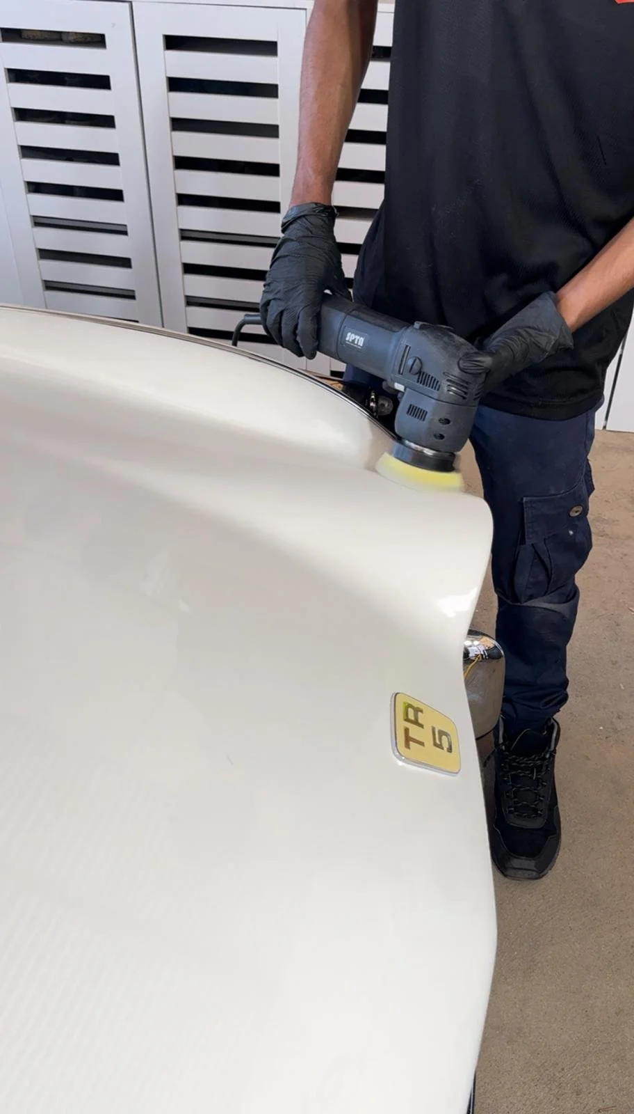 A person polishing a white car using a handheld electric buffer, wearing black gloves and dark clothing.