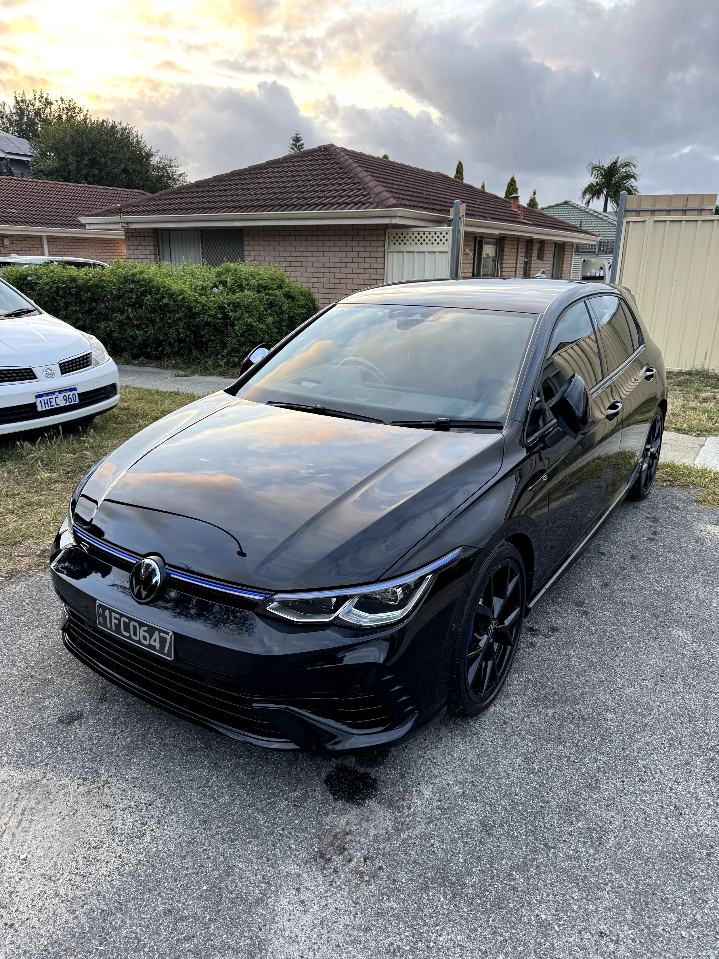 Black Volkswagen car parked in a driveway with a white car visible in the background, houses, bushes, and a cloudy sky
