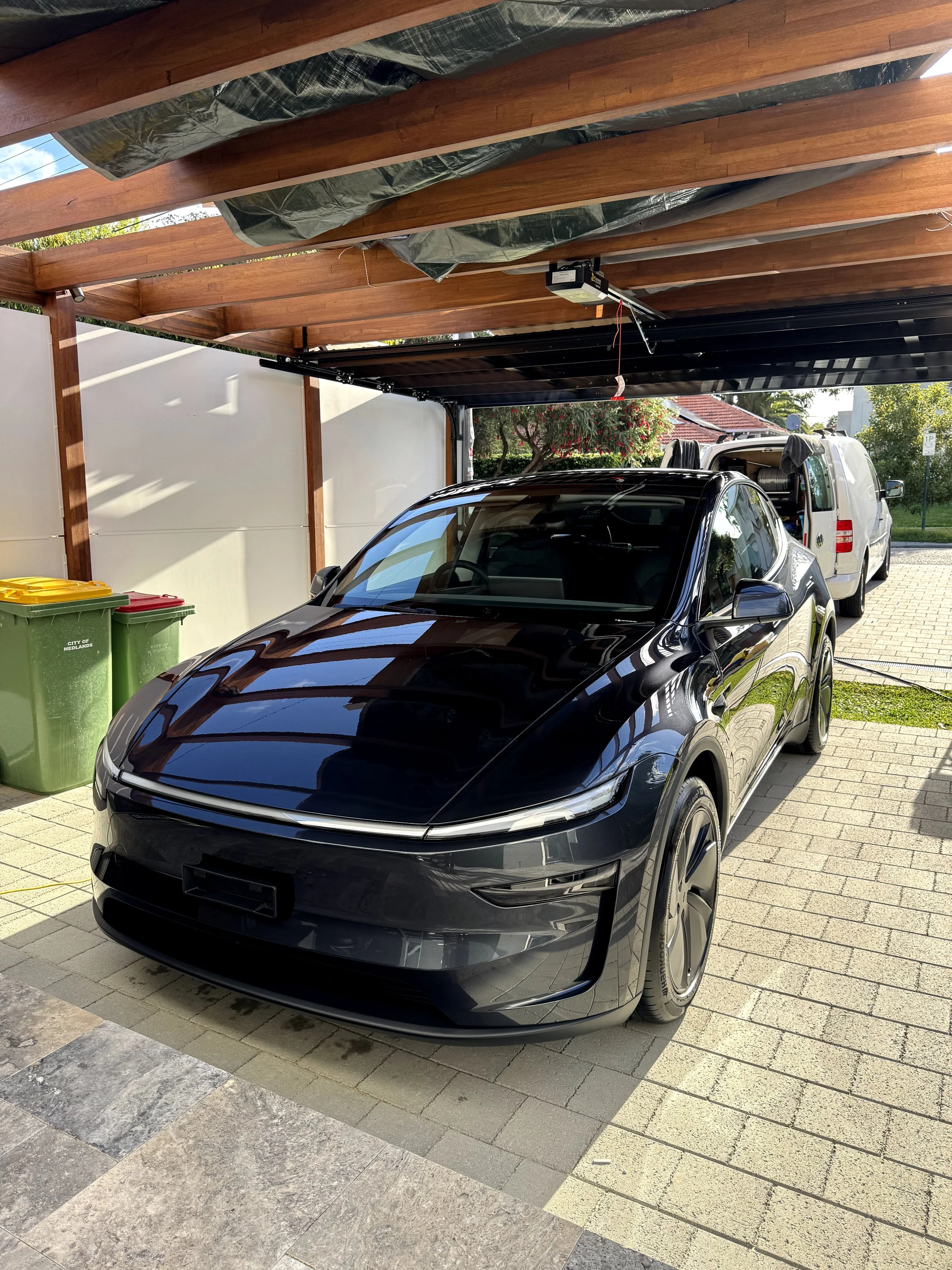 A sleek black electric car, possibly a Porsche, parked under a wooden carport with a partially open roof, next to two green trash bins and a white van in a driveway during daylight.