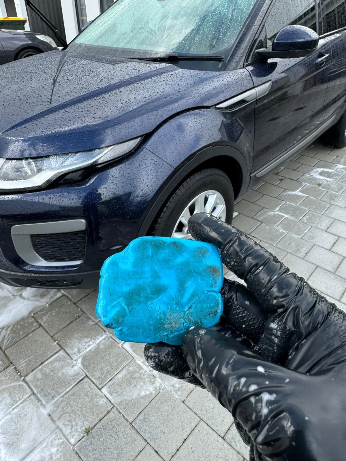 Hand wearing a black glove holding a dirty blue washing sponge in front of a wet black car, on a paved surface with snow.