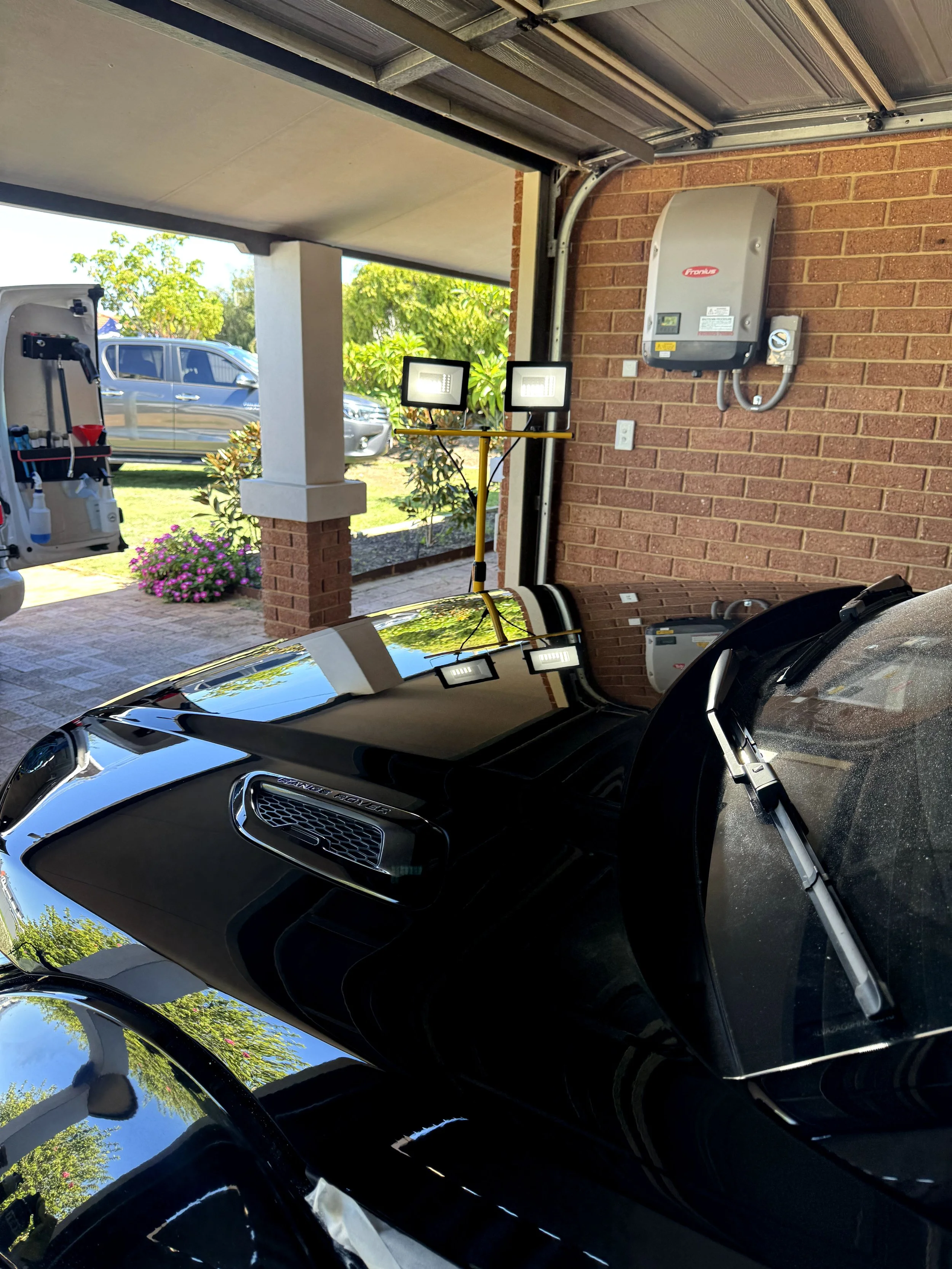 A black sports car parked in a garage, with LED floodlights and an electric vehicle charging station mounted on the brick wall.