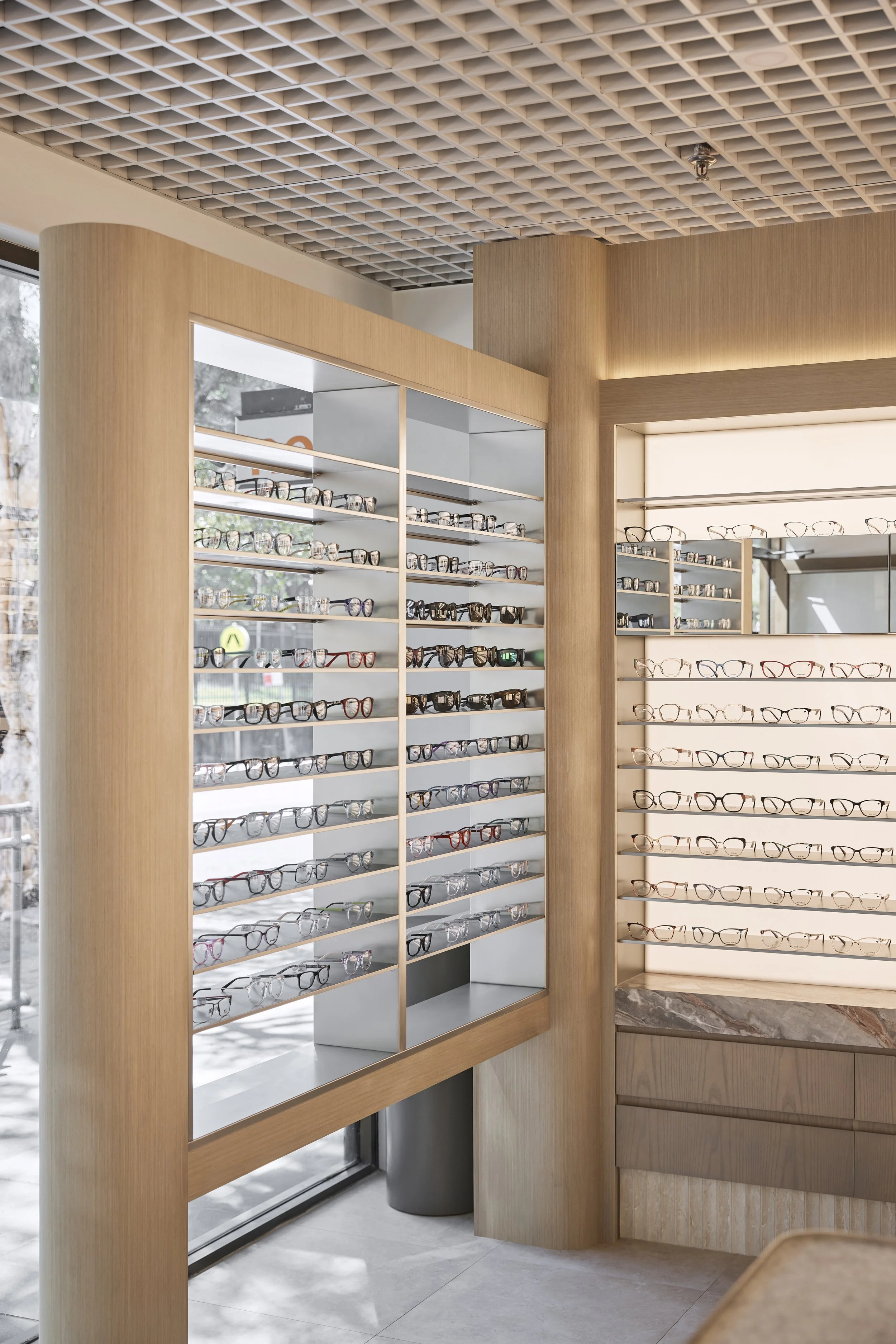 Bright Eyes Optics retail fit-out in Mona Vale, Sydney, featuring custom curved timber joinery, illuminated eyewear displays, detailed stone finishes and feature grid ceiling – Sydney City Renovations.
