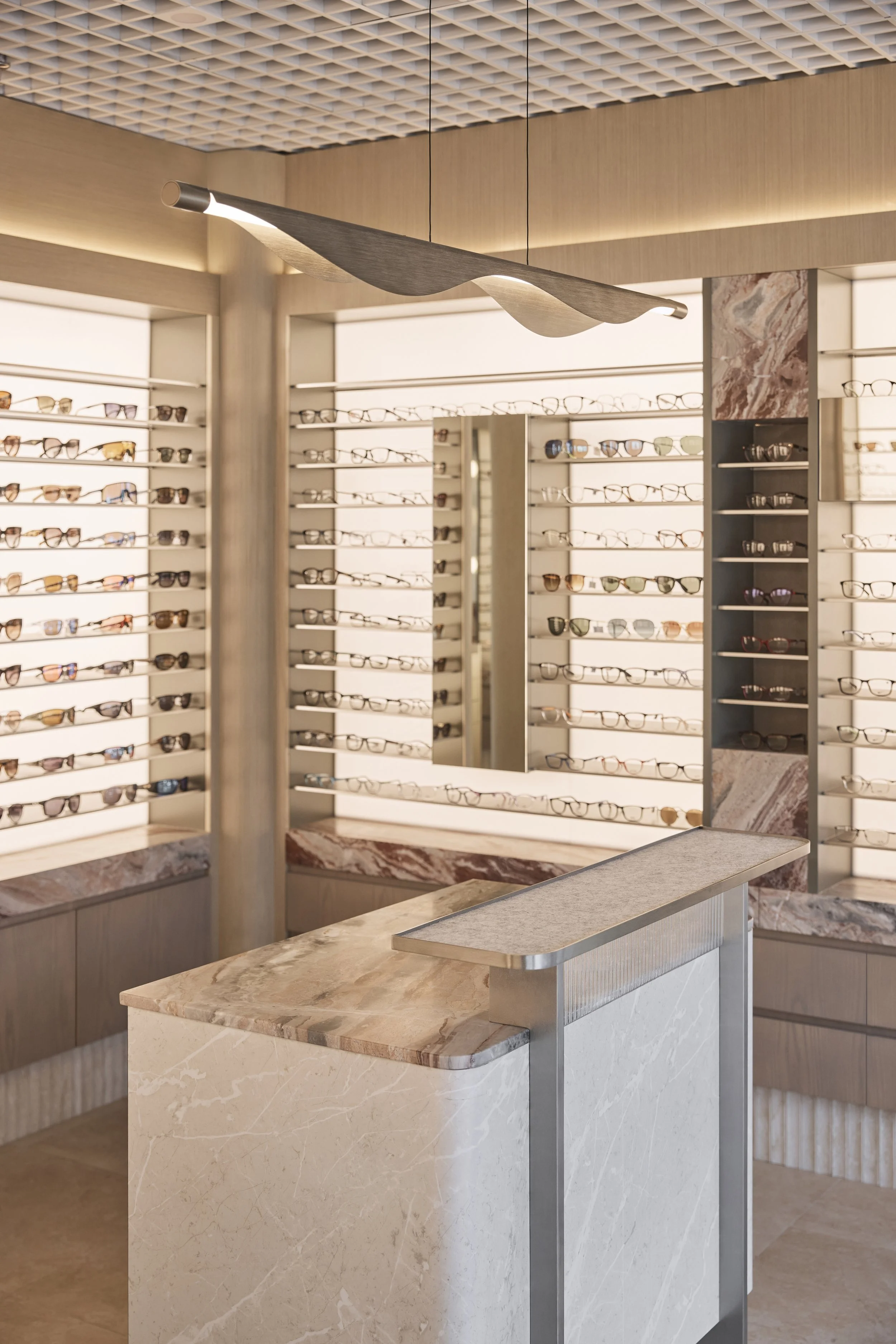 Bright Eyes Optics Mona Vale commercial retail fit-out featuring custom backlit eyewear displays, natural stone reception counter, warm timber joinery, sculptural pendant lighting and decorative grid ceiling – Sydney City Renovations.