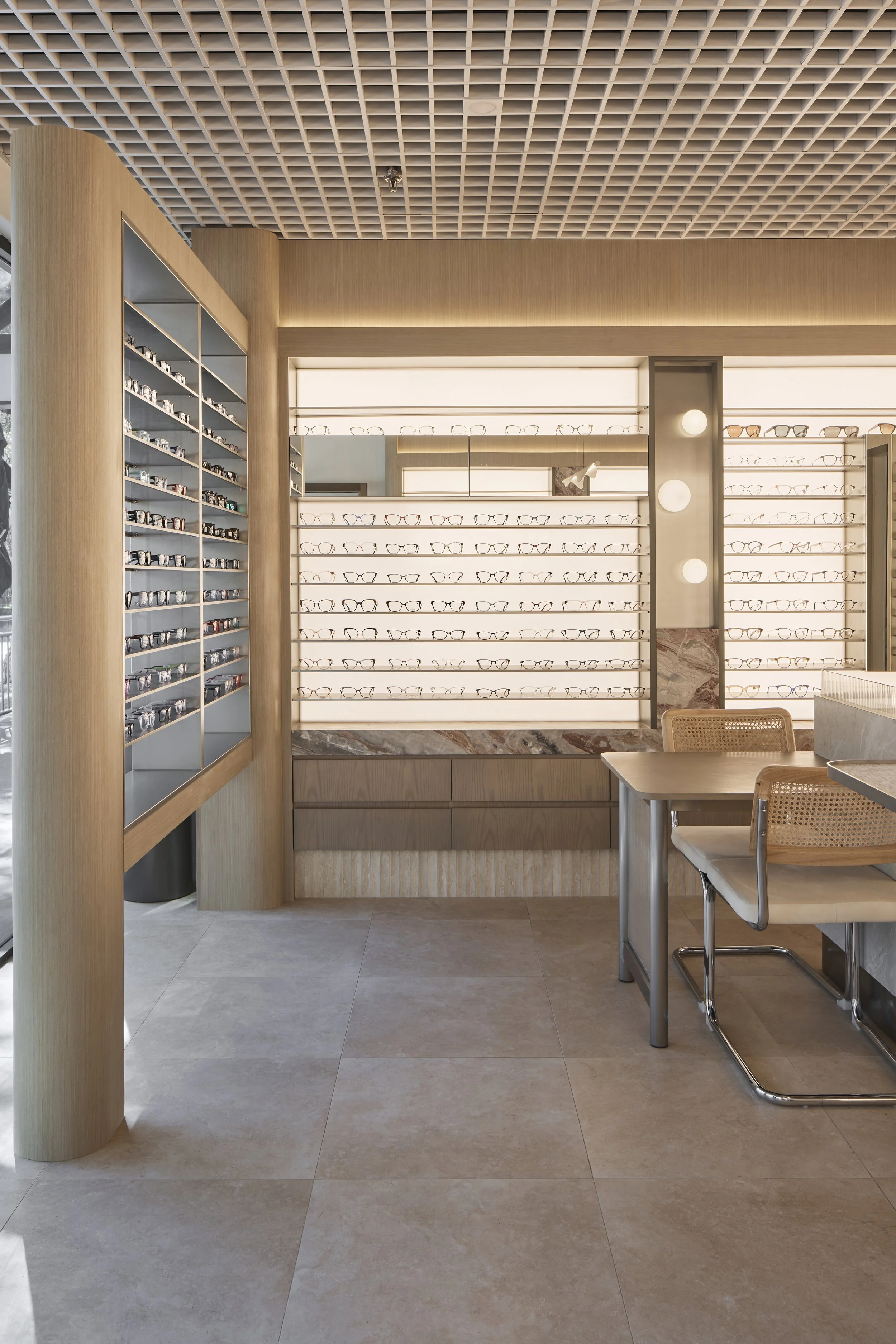 Bright Eyes Optics Mona Vale commercial retail fit-out featuring custom illuminated eyewear displays, warm timber joinery, natural stone finishes, decorative grid ceiling and contemporary consultation furniture – Sydney City Renovations.
