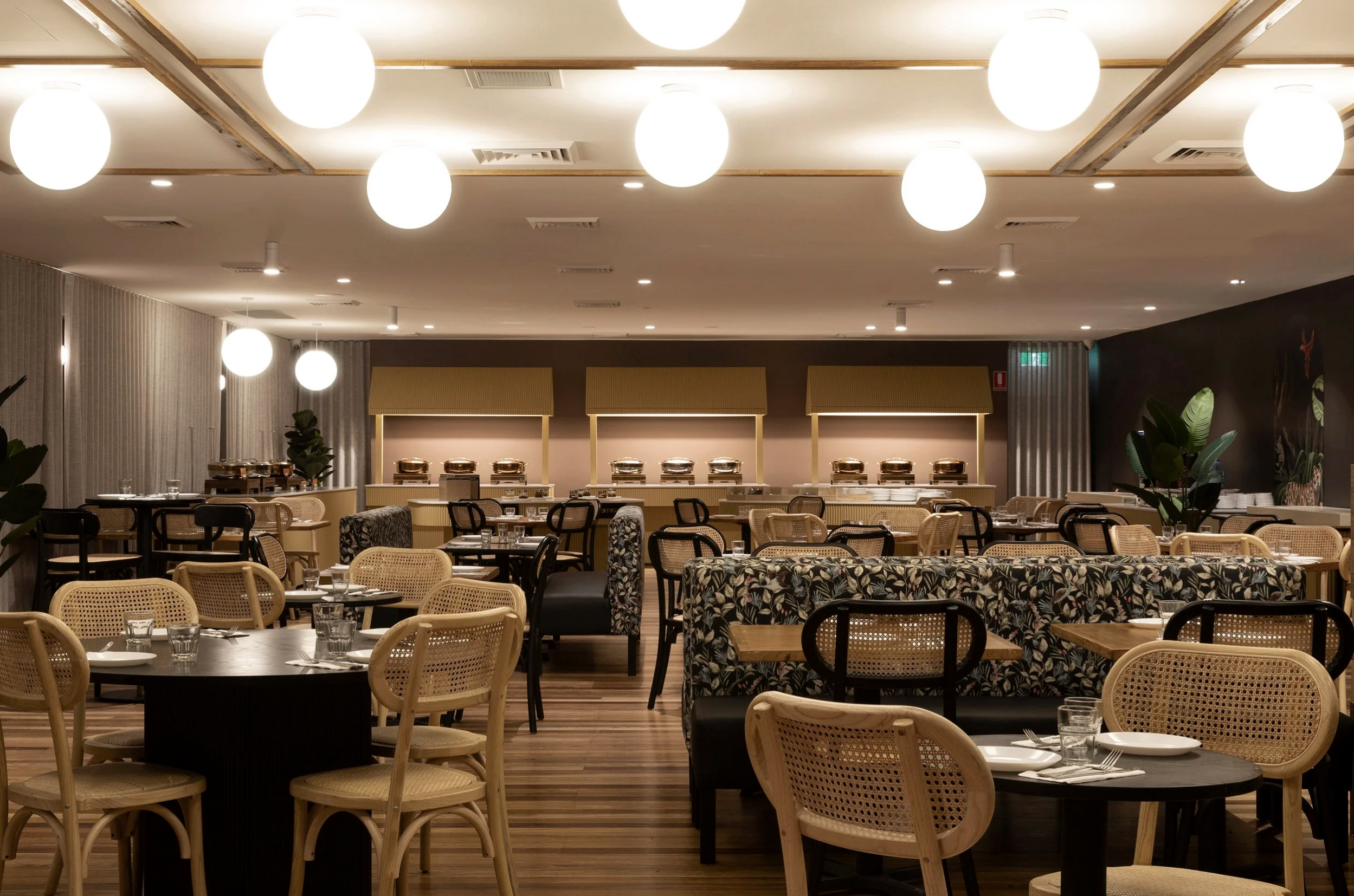 Sydney City Renovations commercial restaurant fitout featuring warm timber flooring, decorative globe lighting, custom floral upholstered banquette seating, natural cane dining chairs and contemporary buffet stations, creating a welcoming and style. 