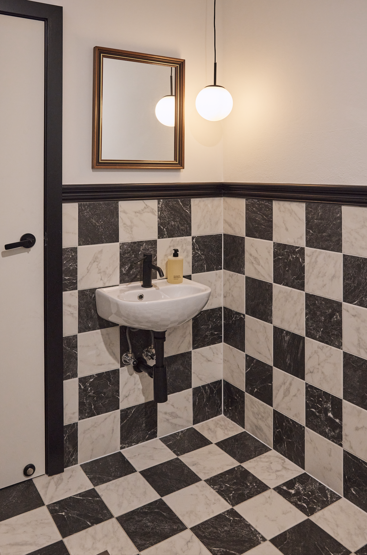 Sydney City Renovations commercial hospitality bathroom fit-out featuring black-and-white marble-look chequered wall and floor tiles, a wall-mounted basin, matte black tapware, decorative timber-framed mirror and contemporary globe pendant lighting.