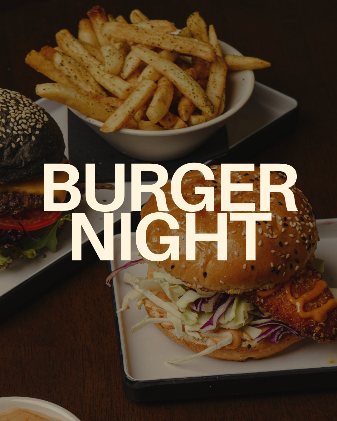 Close-up of a burger with coleslaw in a bun with sesame seeds, side of French fries in a bowl, and another burger with lettuce and tomato on a plate, on a dark table.