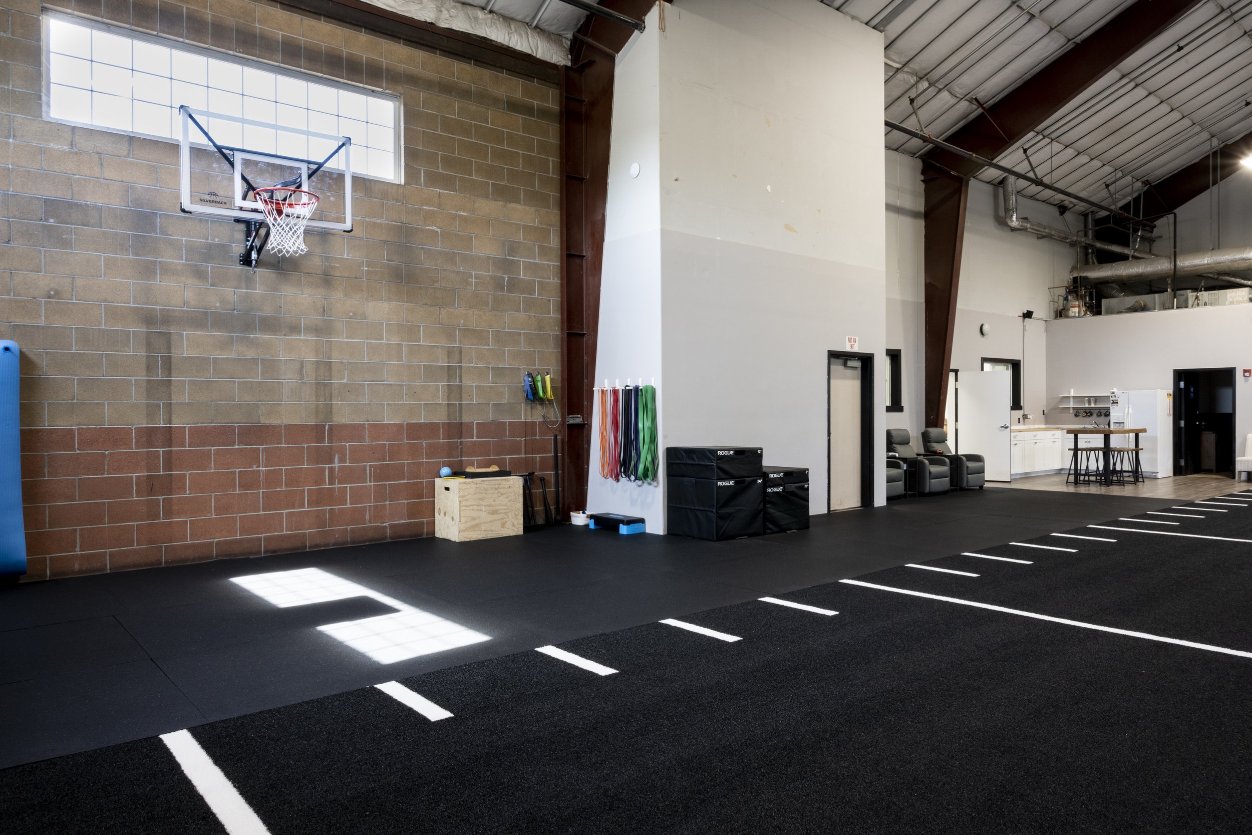Indoor gym space with basketball hoop, resistance bands, seating area, and kitchenette. Features high ceilings and black rubber flooring with white lines.