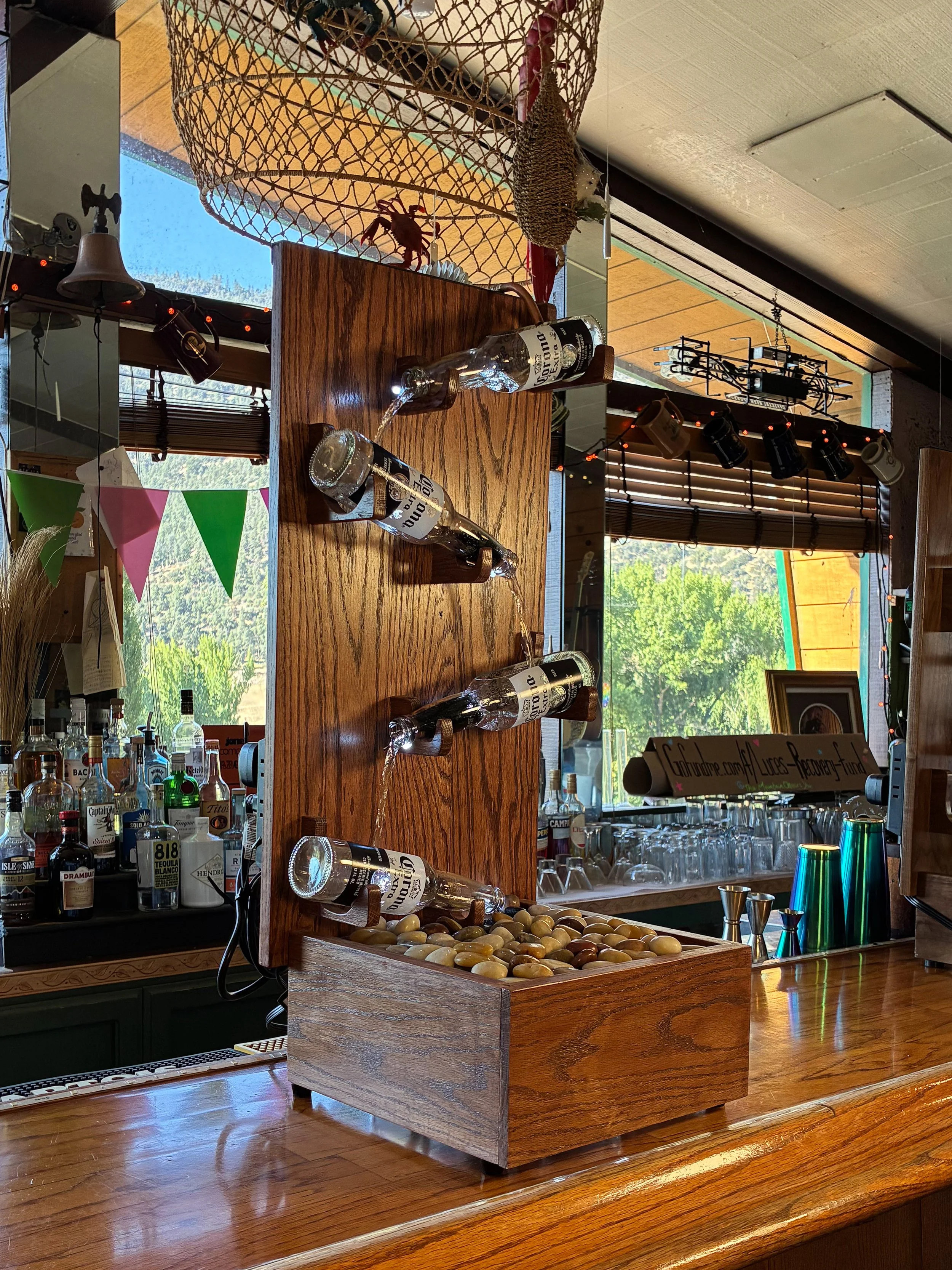 Angled view of a red oak beer bottle fountain showing the wooden backboard, bottle arrangement, and river rock basin.
