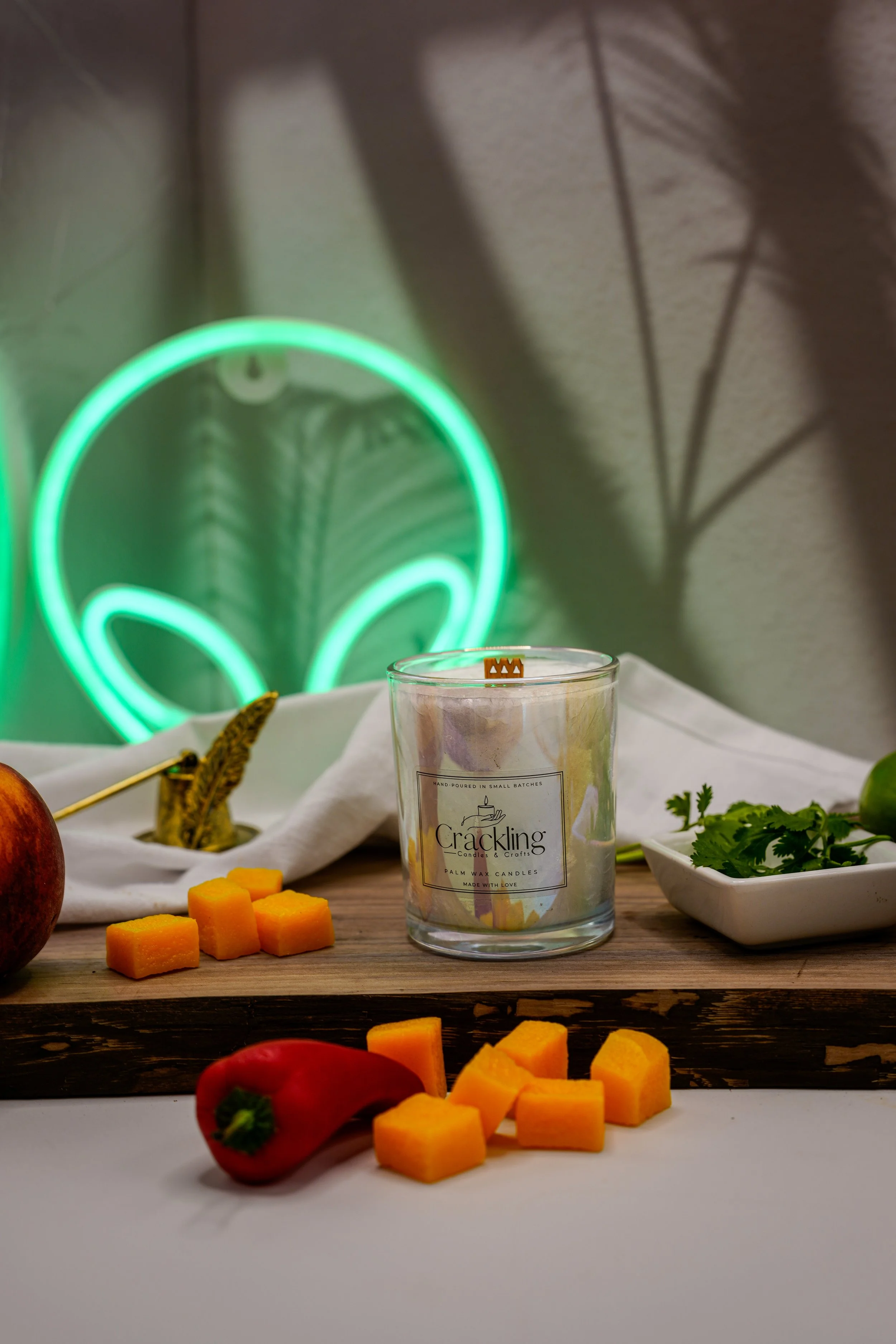 Fireball candle in a glass jar on a wooden board, styled with cream fabric, colorful peppers, and a glowing green alien sign in the background.