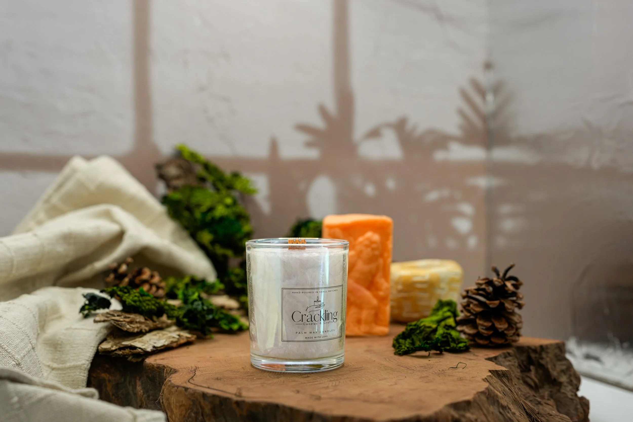 Cardamom & Teakwood candle displayed on natural wood with greenery, pinecones, soft linen, and warm amber accents.