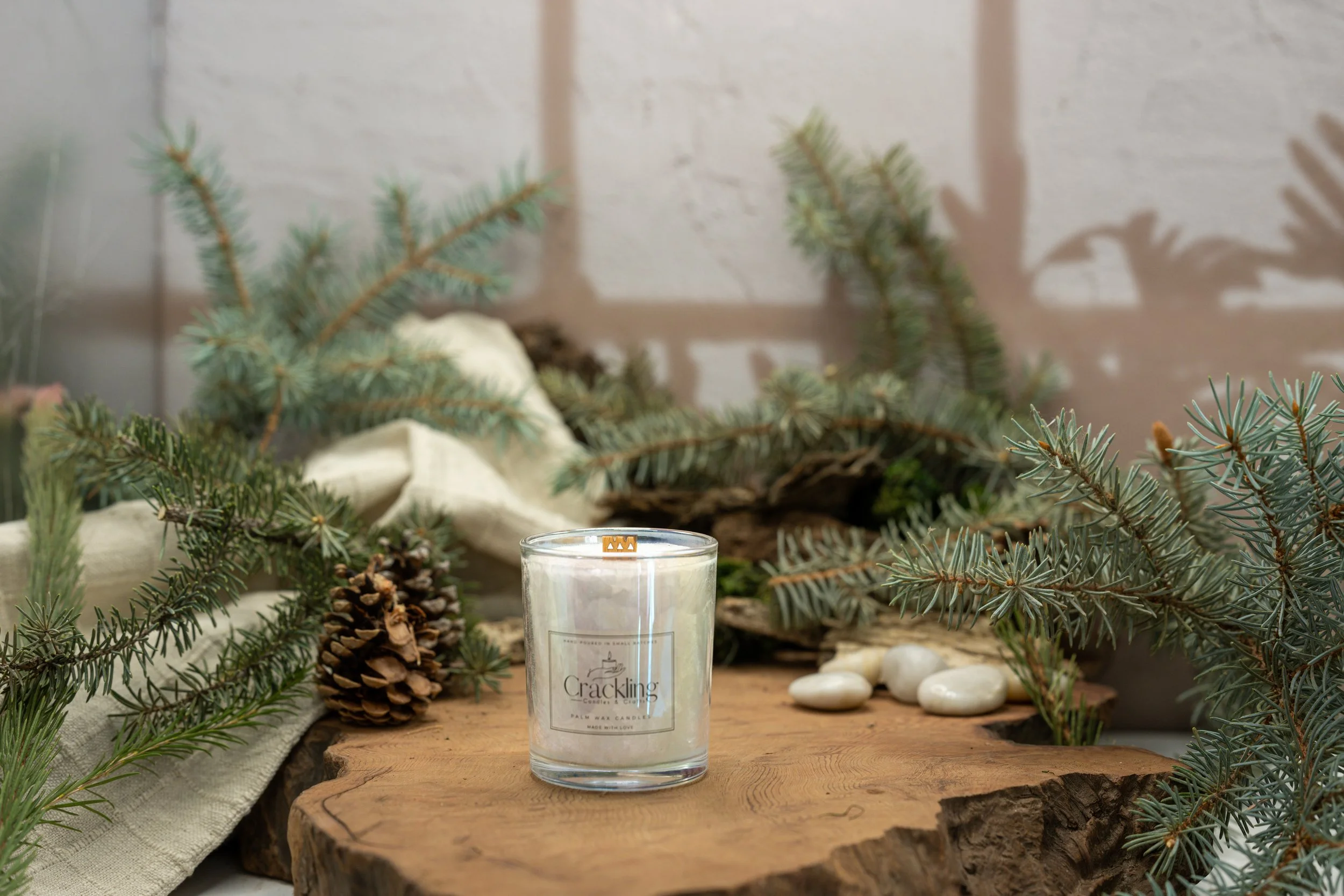 Forest Pine candle in a glass jar on a live-edge wood slab, styled with evergreen branches, pinecones, cream fabric, and smooth stones.