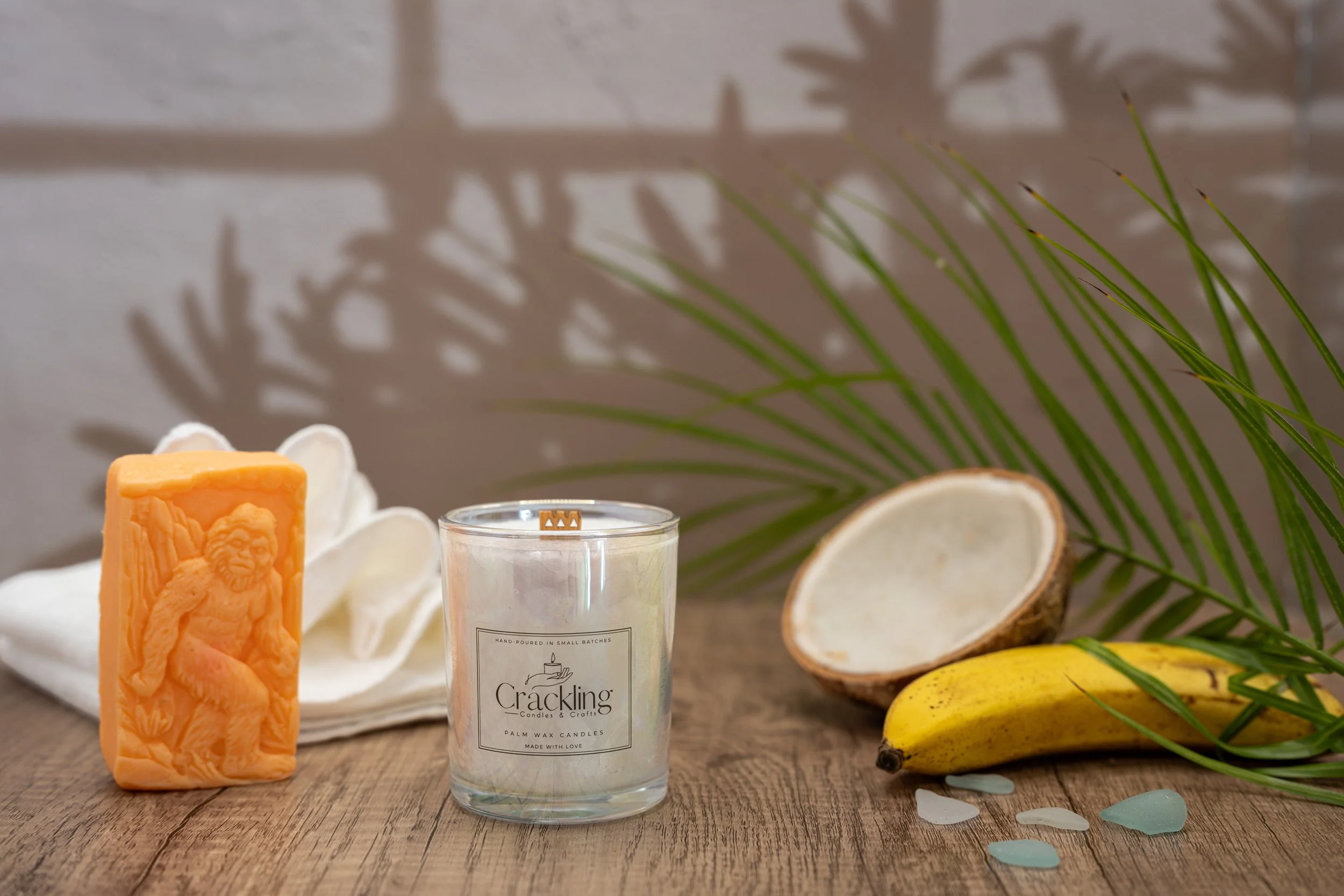Front view of the 6 oz Butt Naked palm wax candle with its label visible, styled beside coconut, banana, and palm leaves.