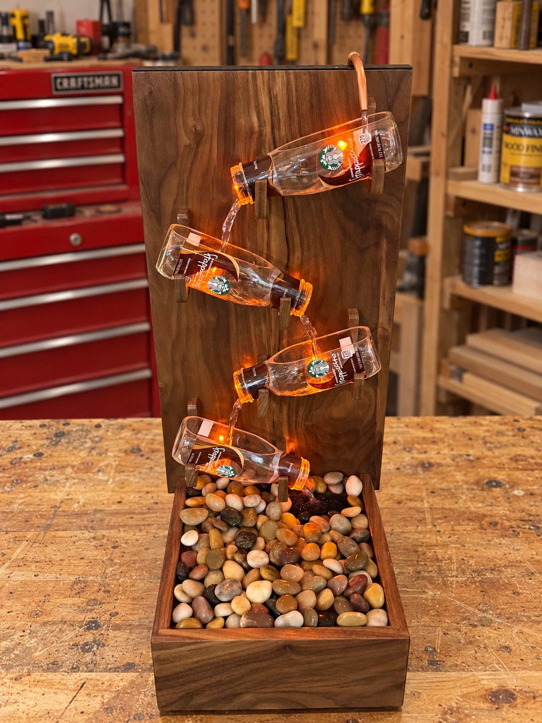 Black walnut fountain on a workshop table with four illuminated Frappuccino bottles above a river rock basin.