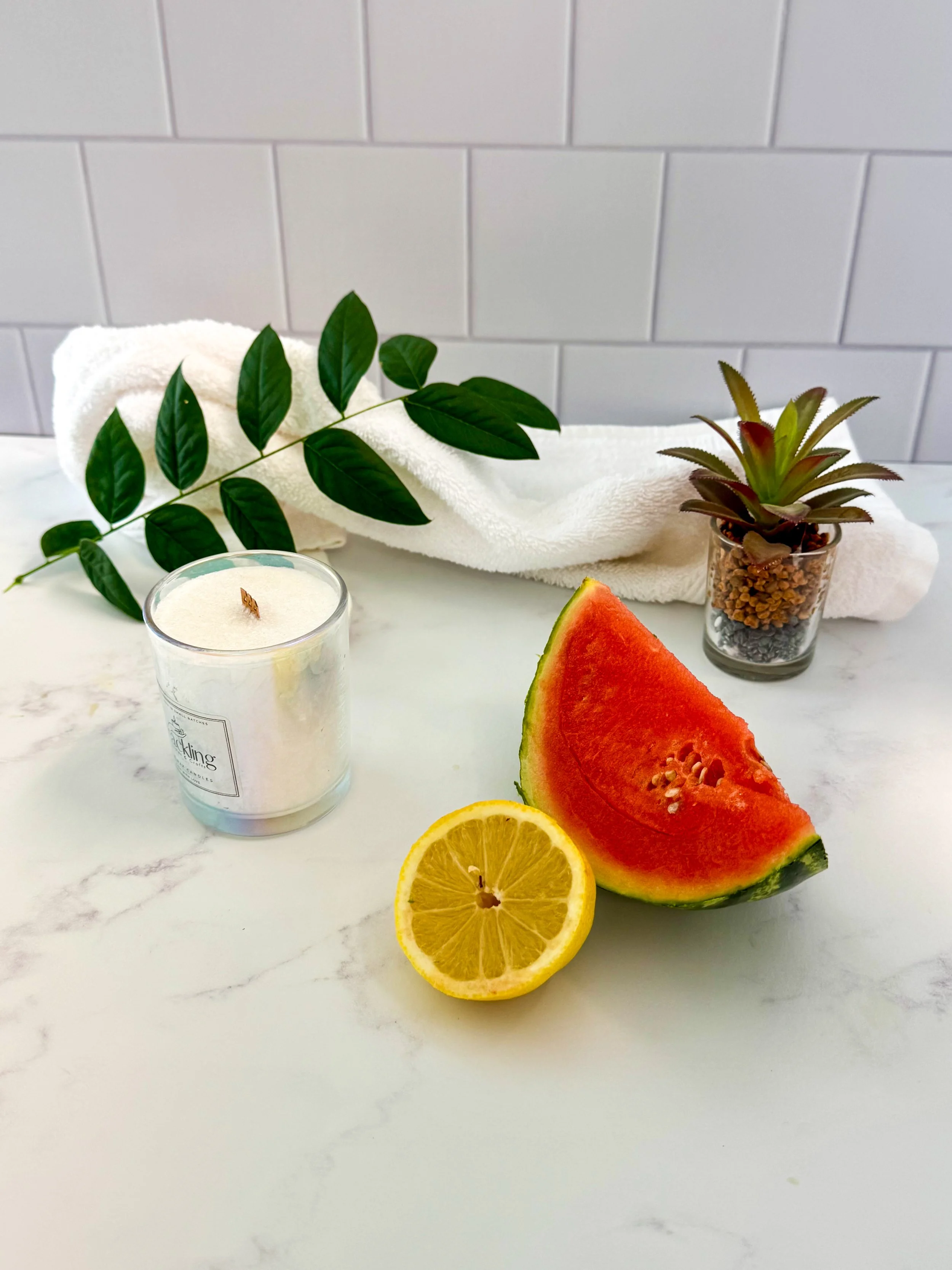 Palm wax candle styled with watermelon, a lemon half, green leaves, and a small potted plant.