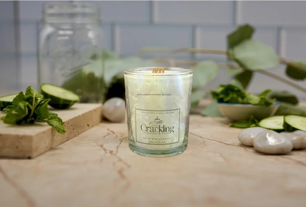 Palm wax candle in an iridescent glass jar, styled with cucumber slices and green leaves on a light countertop.