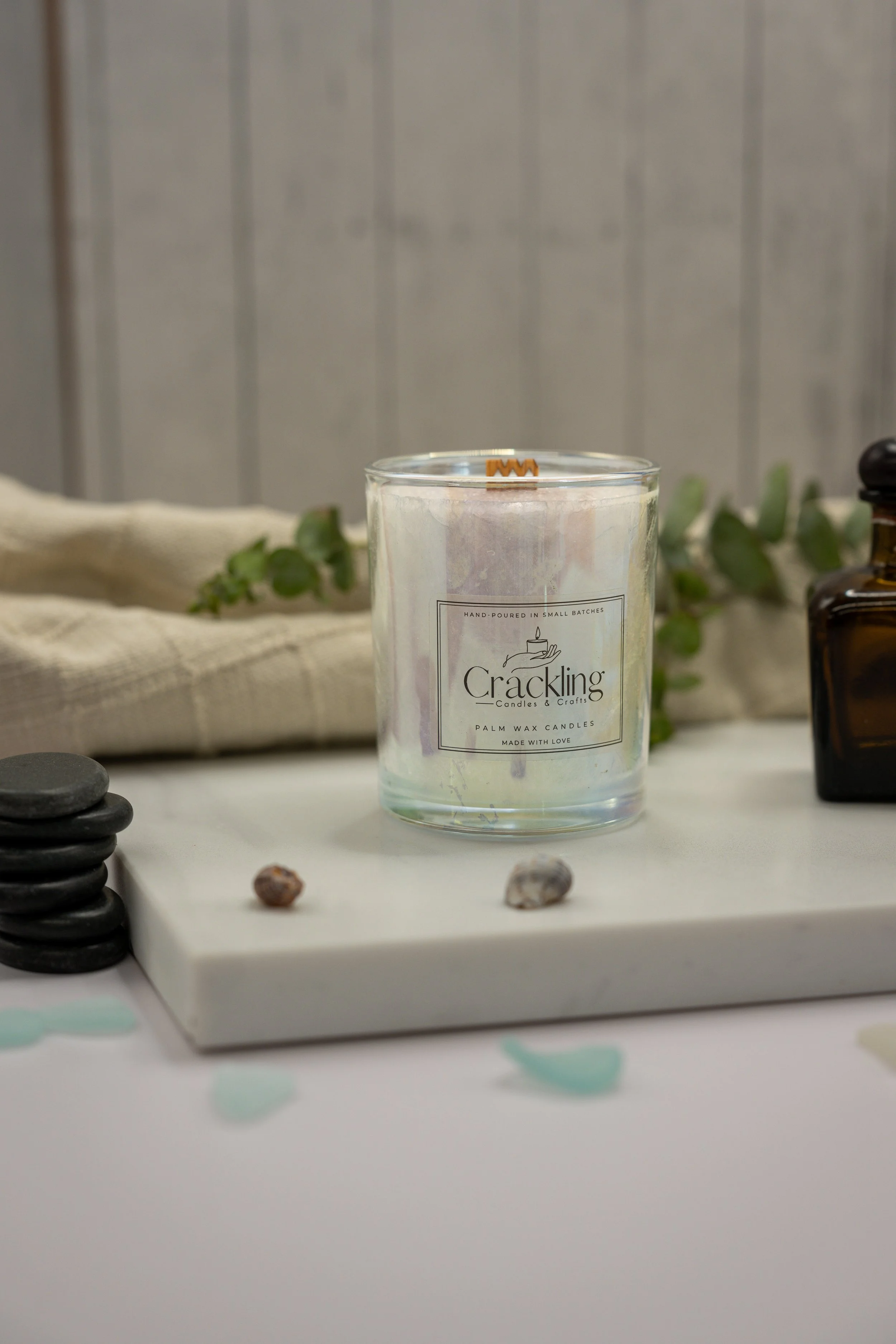 Lemongrass palm wax candle in a clear glass jar on a white slab, styled with stones, greenery, cream fabric, and an amber bottle.