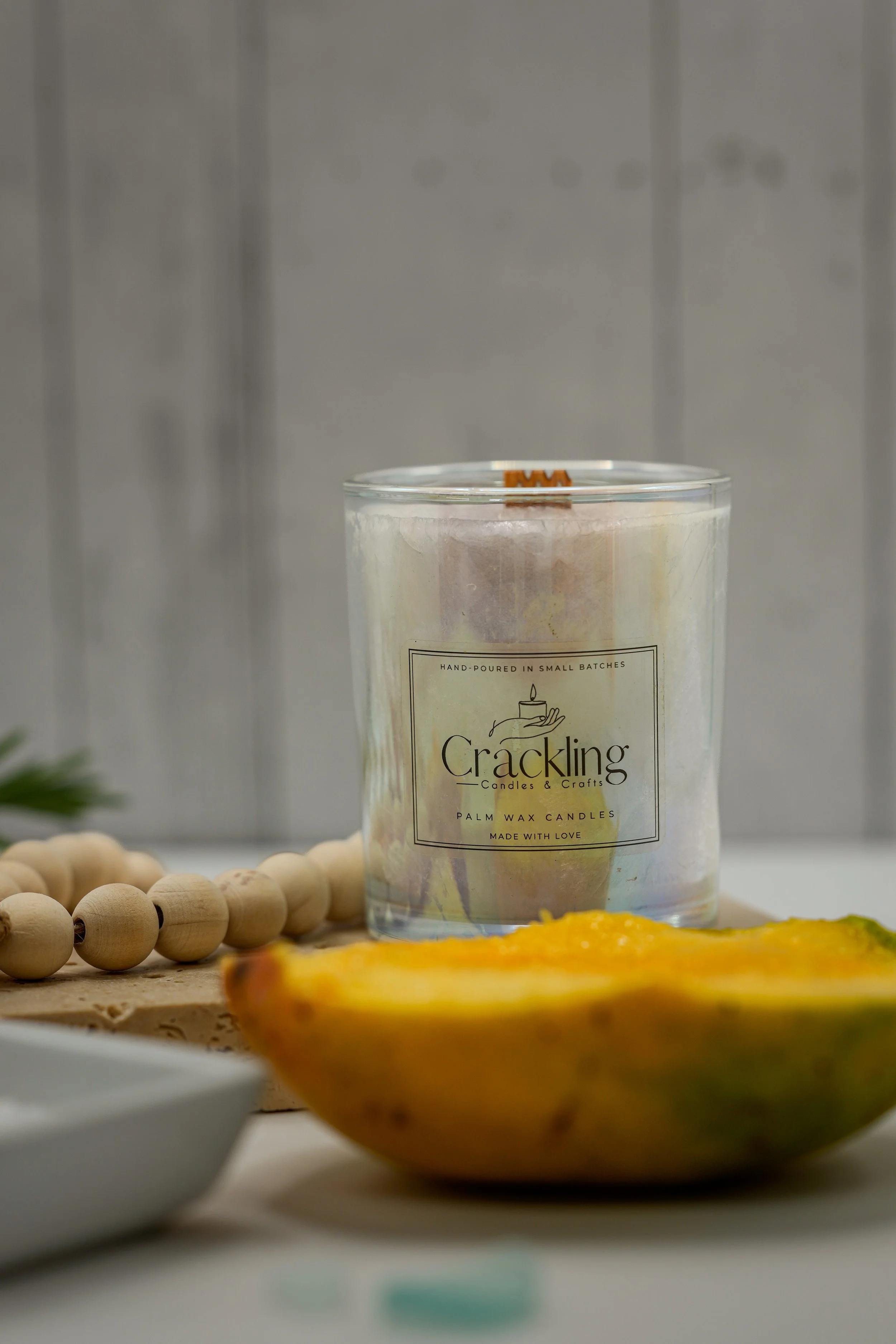 Glass jar palm wax candle with the Crackling Candles & Crafts label, styled with a papaya half and wooden beads.