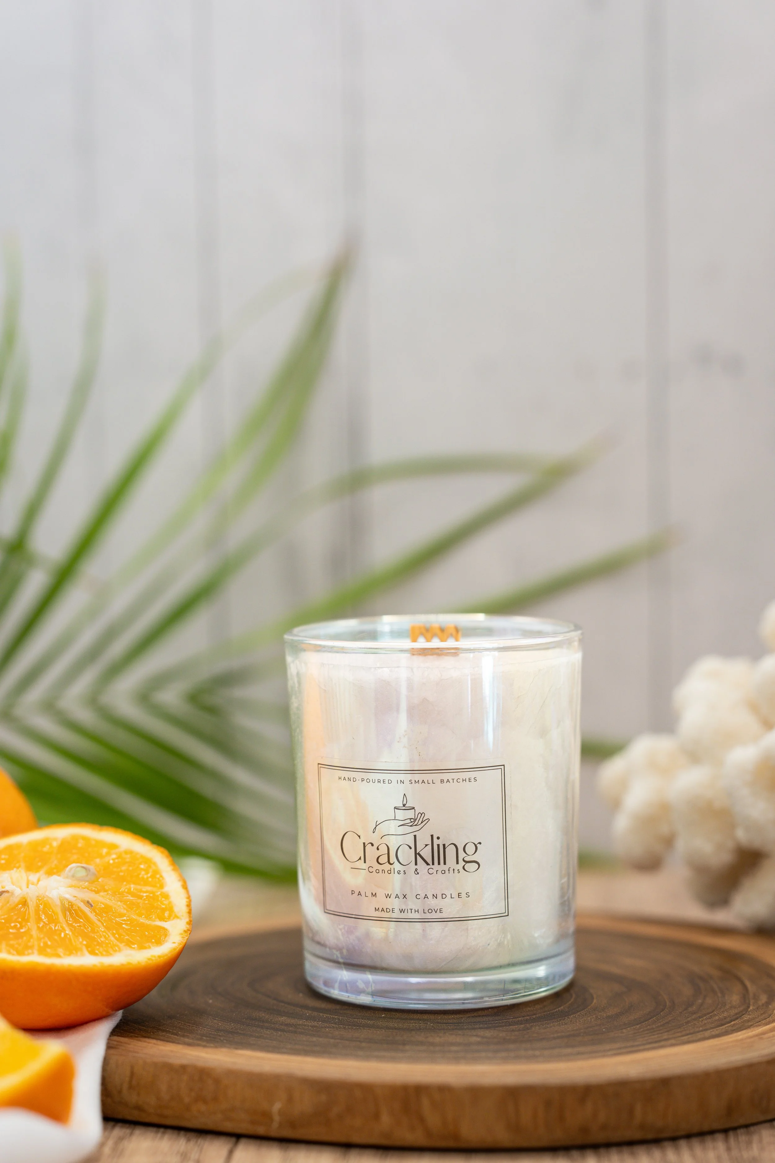 Orange Goji Berry palm wax candle in a clear glass jar, styled with an orange half, green palm leaves, and cream fabric.