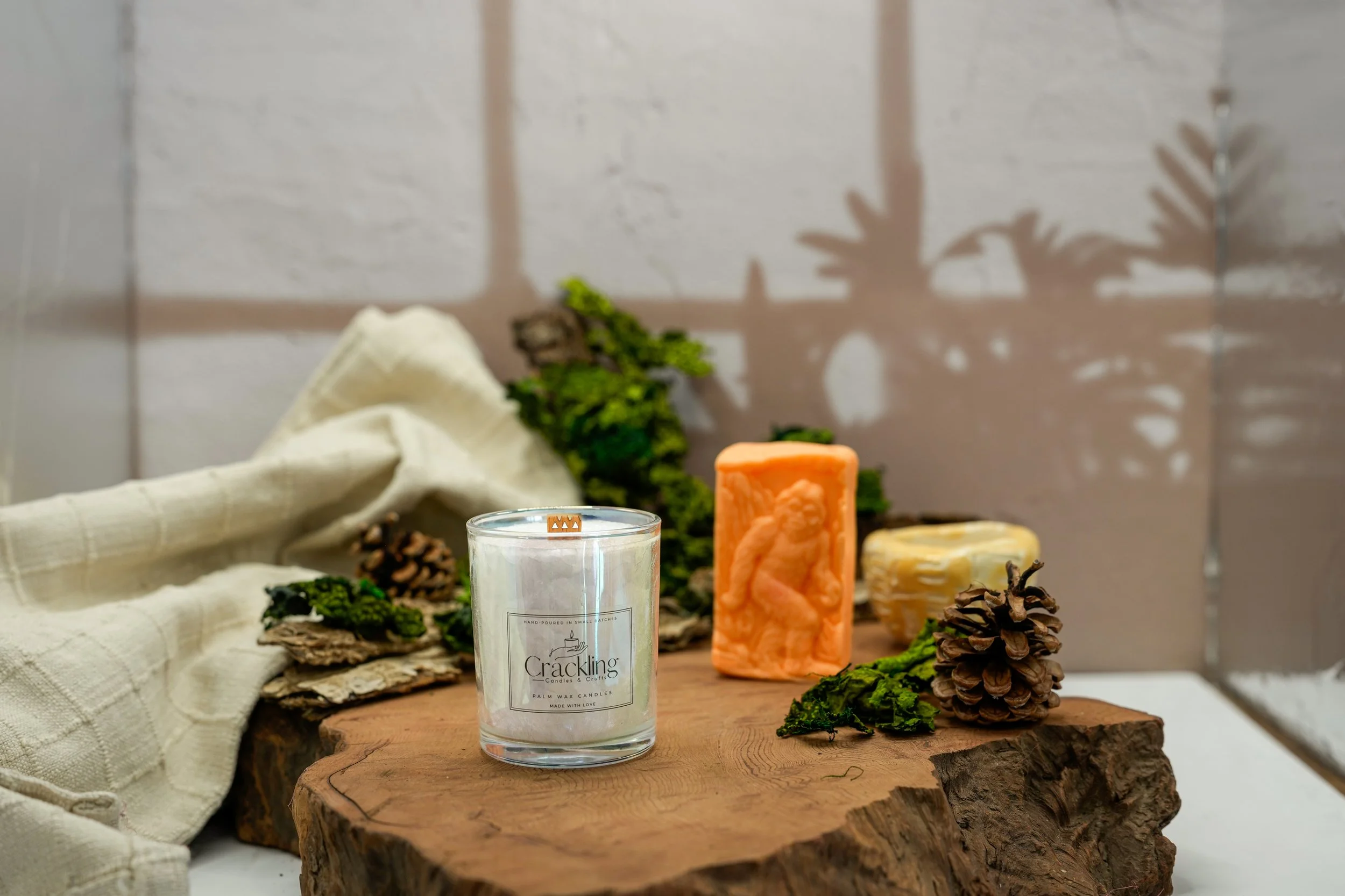 Hand-poured Cardamom & Teakwood candle styled with evergreen details, pinecones, cream linen, and rustic wood.
