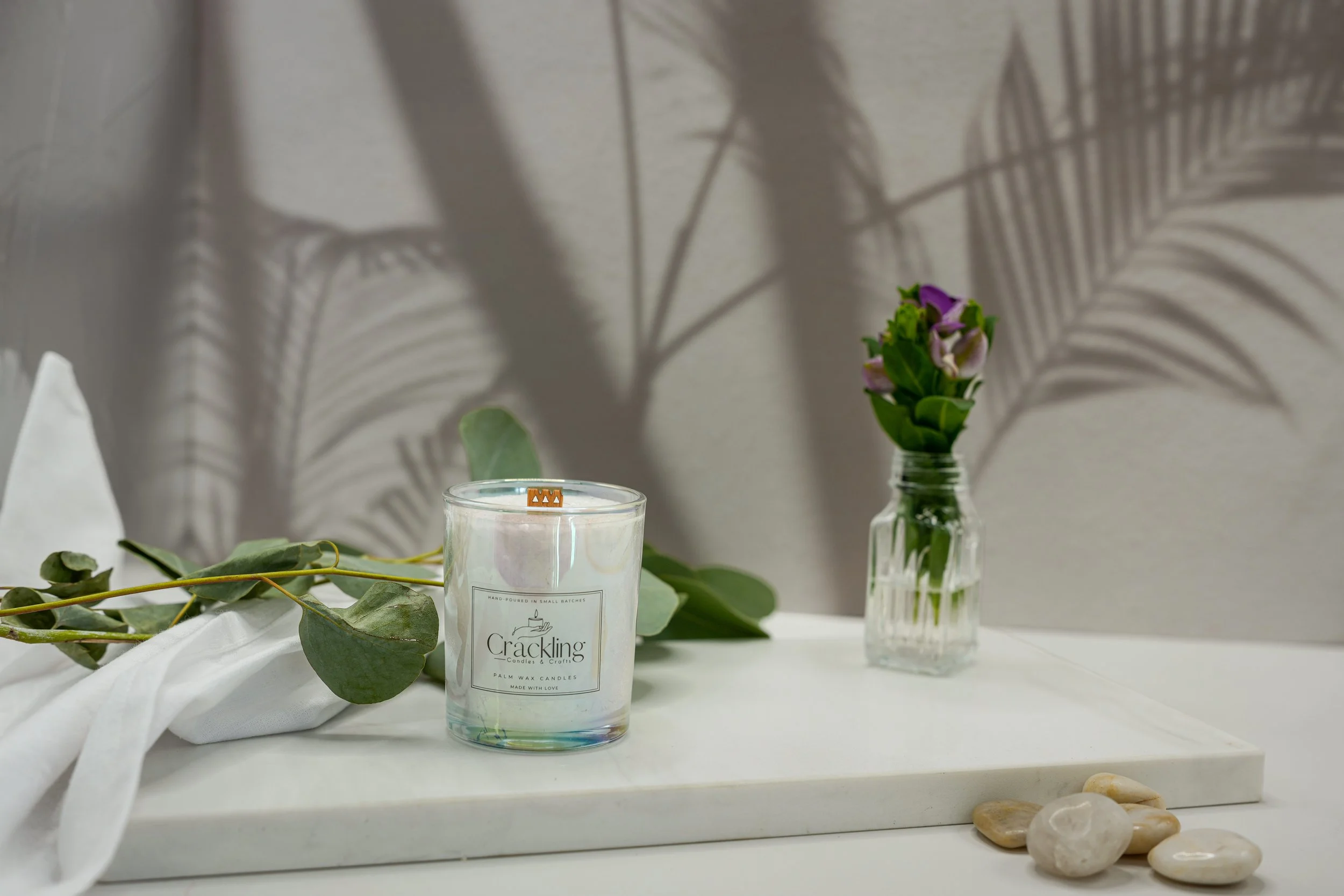 6 oz Indian Sandalwood palm wax candle in an iridescent glass holder, styled on a white surface with greenery and smooth stones