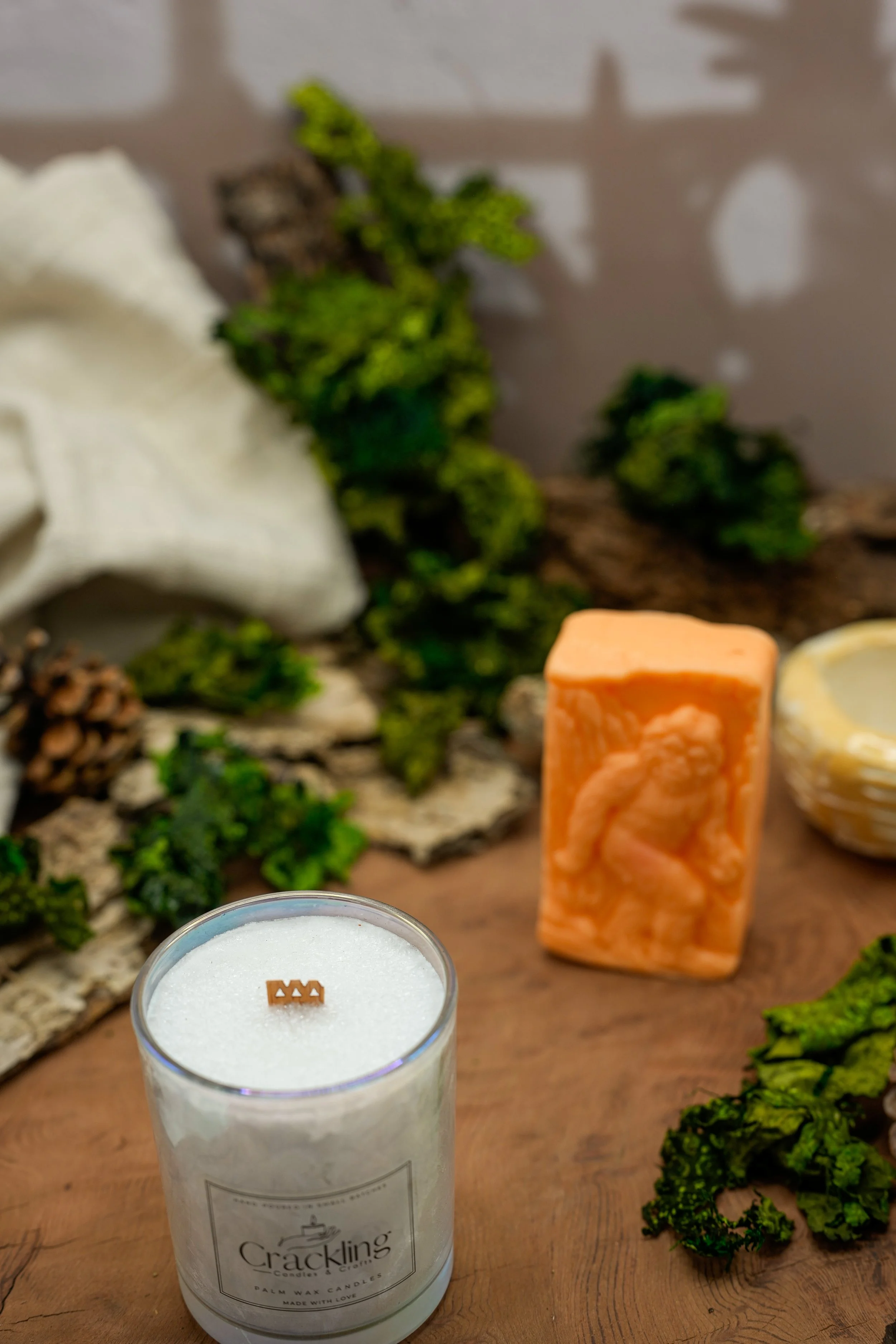 Close-up of a Cardamom & Teakwood palm wax candle with a wooden wick, greenery, pinecones, and warm amber decor.