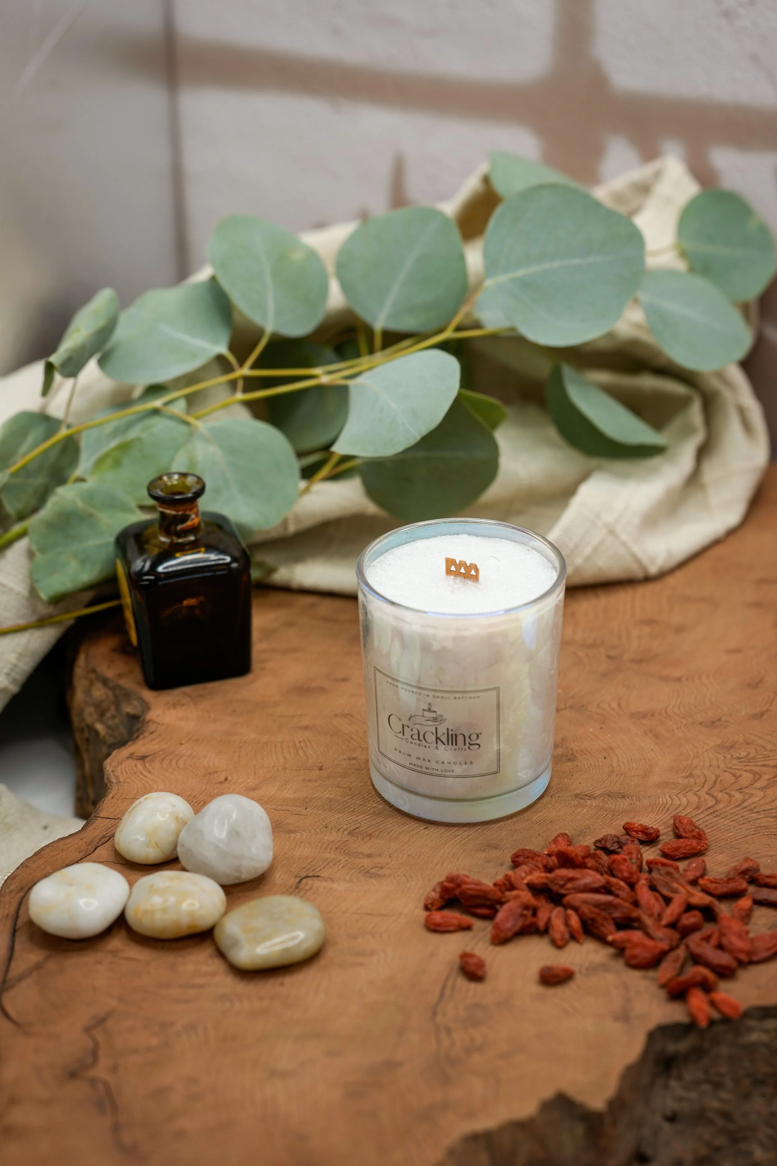 Palm wax candle with a wooden wick beside green leaves, pale stones, a small amber bottle, and dried botanicals.