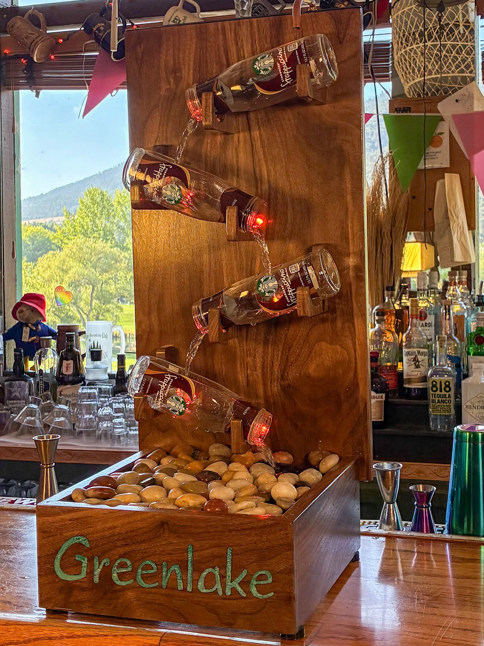 Angled view of a black walnut fountain showing the cascading Frappuccino bottles, wood grain, and river rock basin.