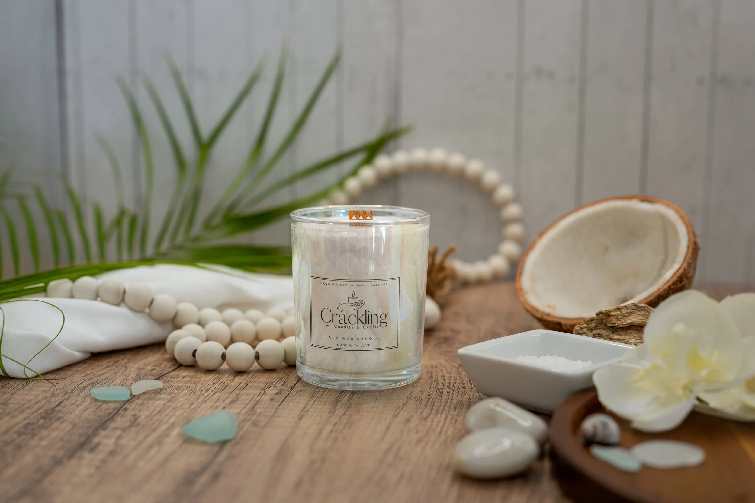 Coconut candle displayed with palm greenery, coconut shells, white beads, and smooth stones on rustic wood.