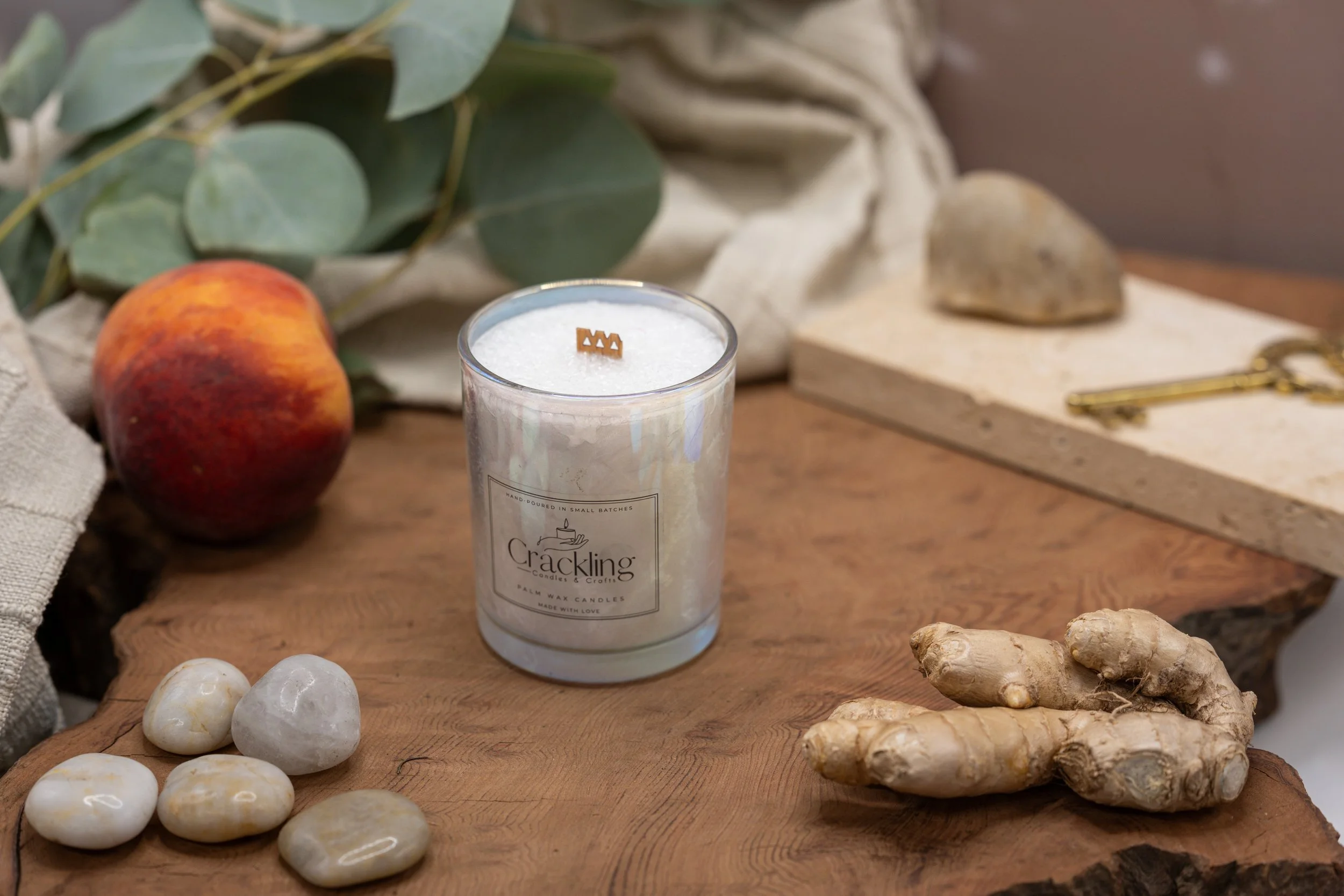 Palm wax candle in an iridescent glass jar on a wooden surface beside ginger root, pale stones, and green leaves.