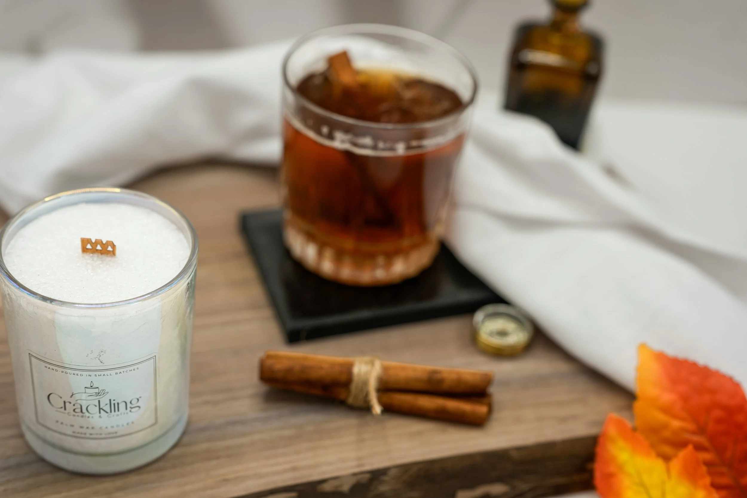 Angled view of an amber drink and bundled cinnamon sticks with a Maple Whiskey Toddy candle partially visible at the left.