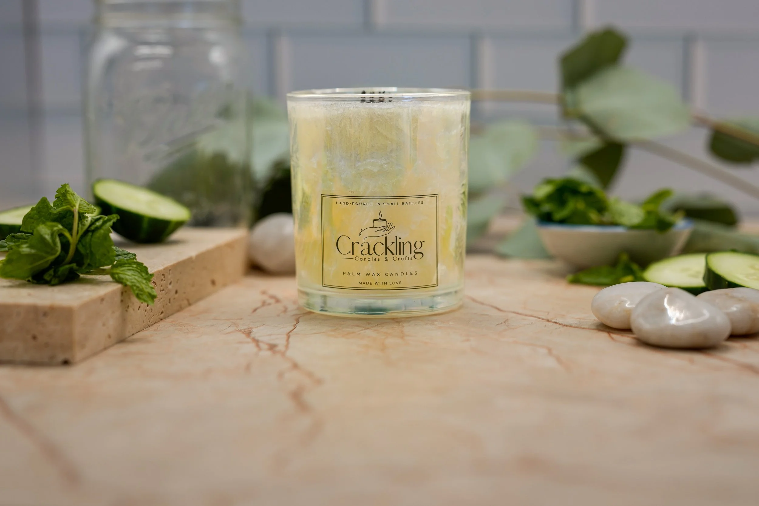 Garden Mint & Cucumber candle in a glass jar shown in a wider view on a beige marble-patterned surface, with cucumber slices and greenery.