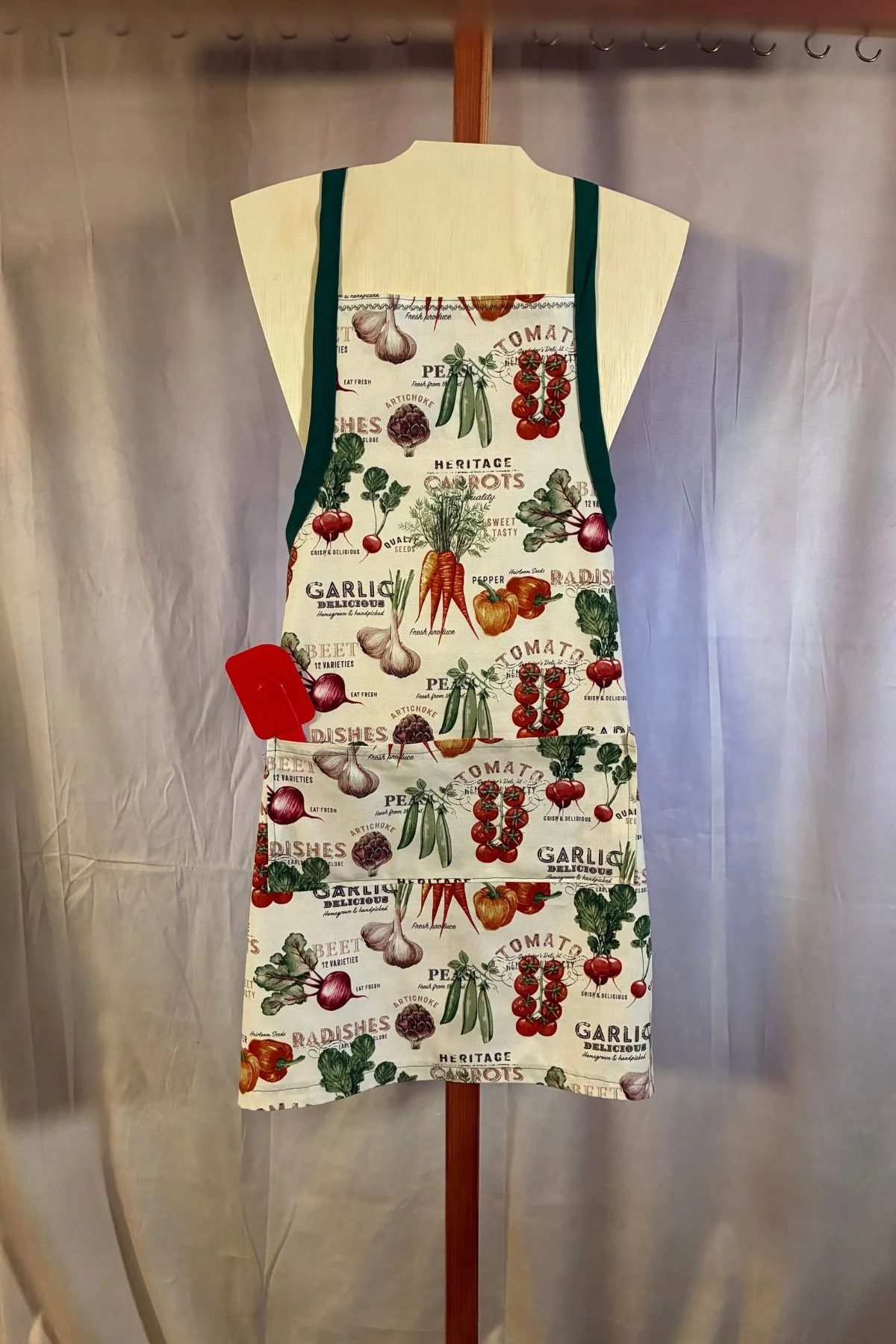 Close view of the vegetable-print cotton apron, showing dark edging and a front pocket with a red spatula displayed inside.