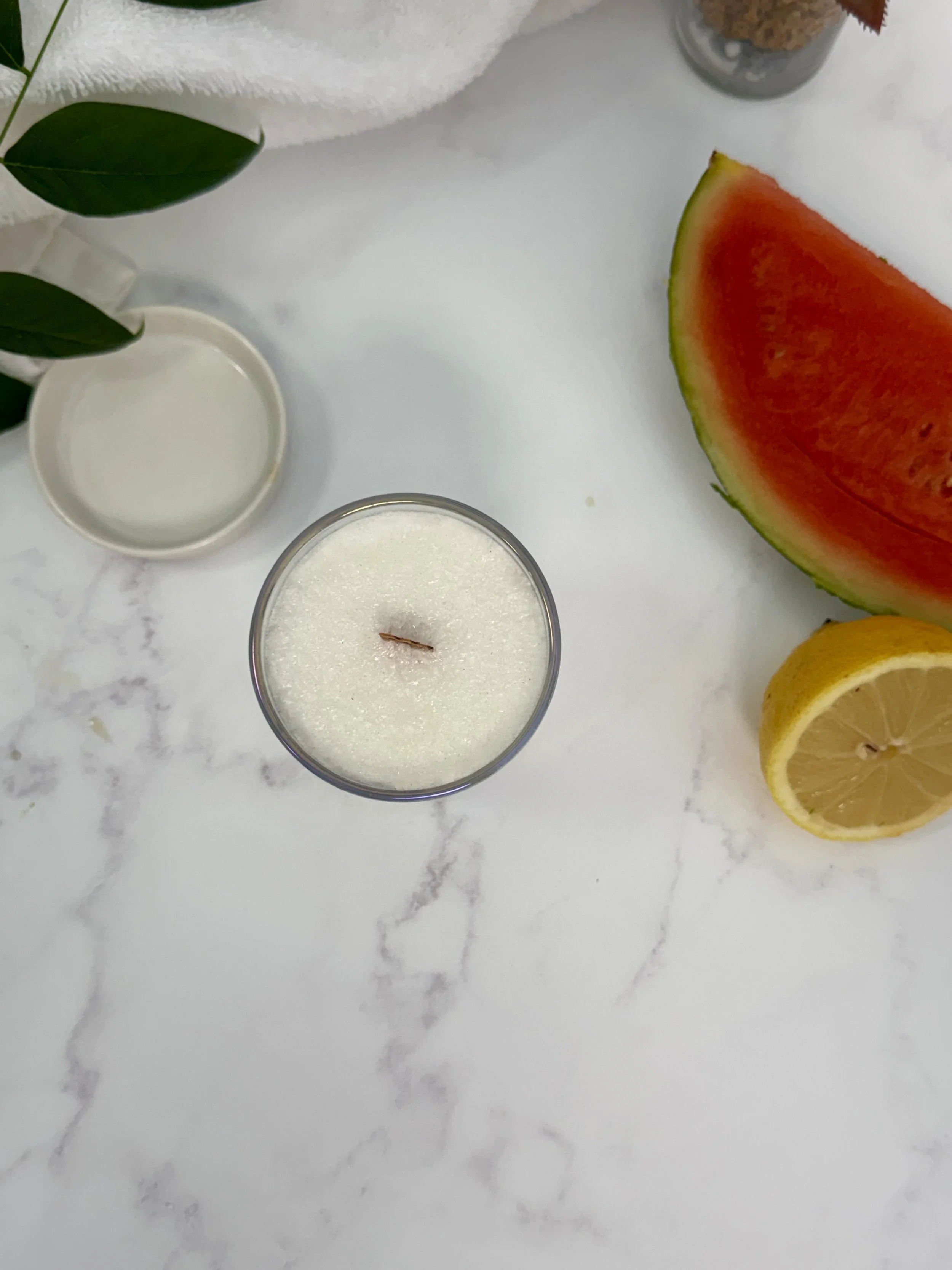Overhead view of a palm wax candle with a wooden wick beside watermelon and a lemon half.