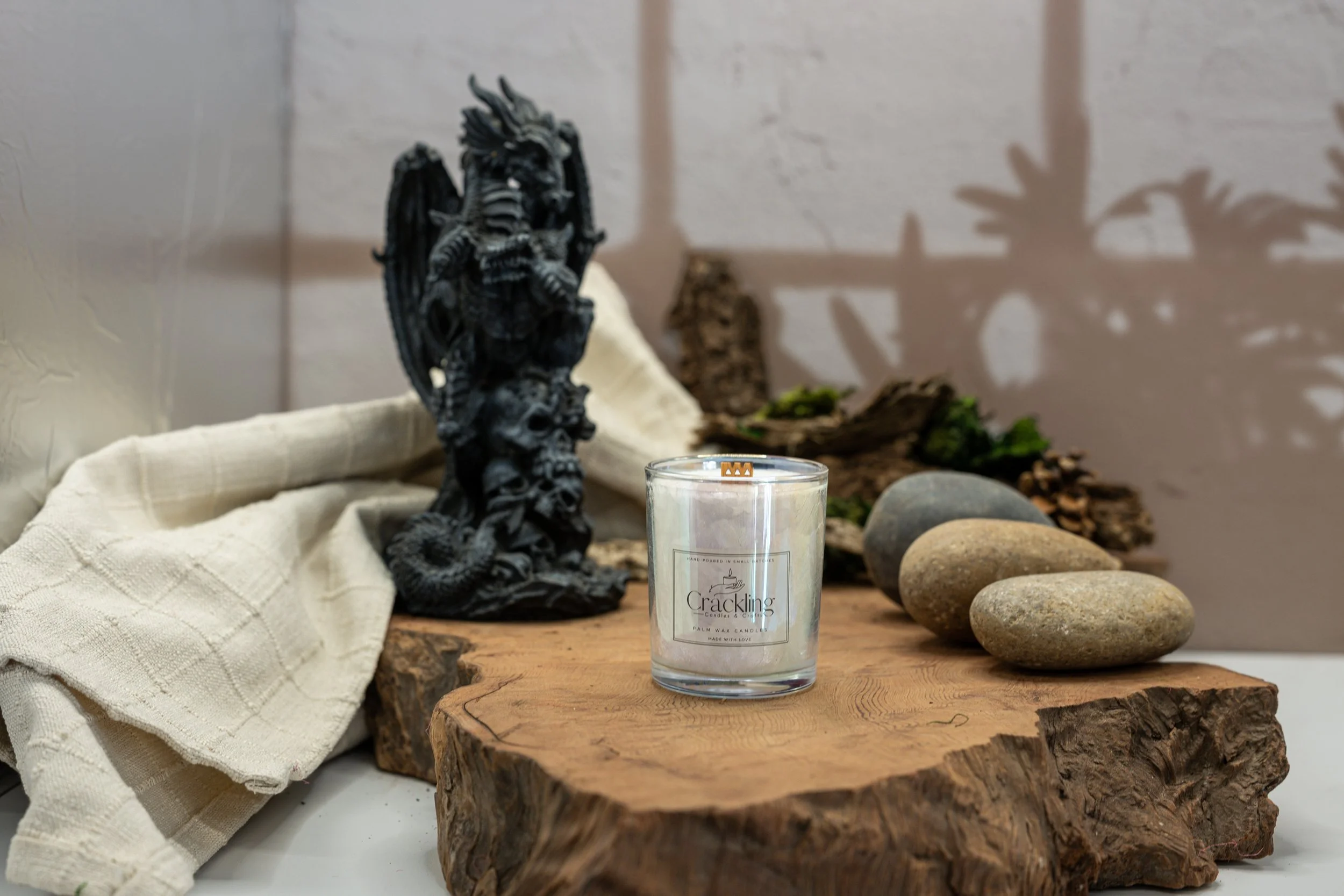 Dragon’s Blood candle in a clear glass jar on a live-edge wood slab, beside a black dragon figurine, smooth stones, and cream fabric.