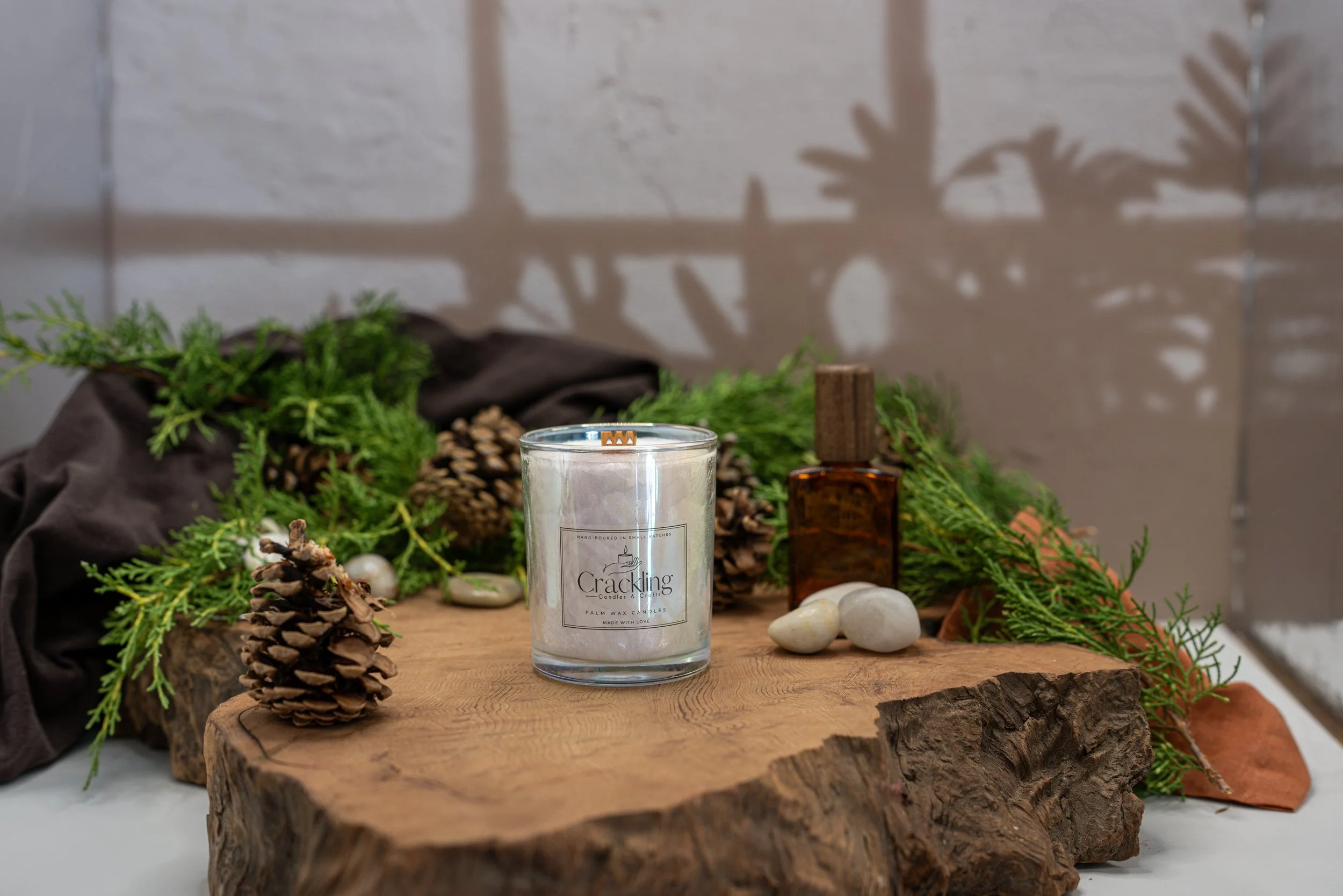 Cedarwood and Amber candle displayed on a rustic wood slab with greenery, pinecones, white stones, and warm amber accents.