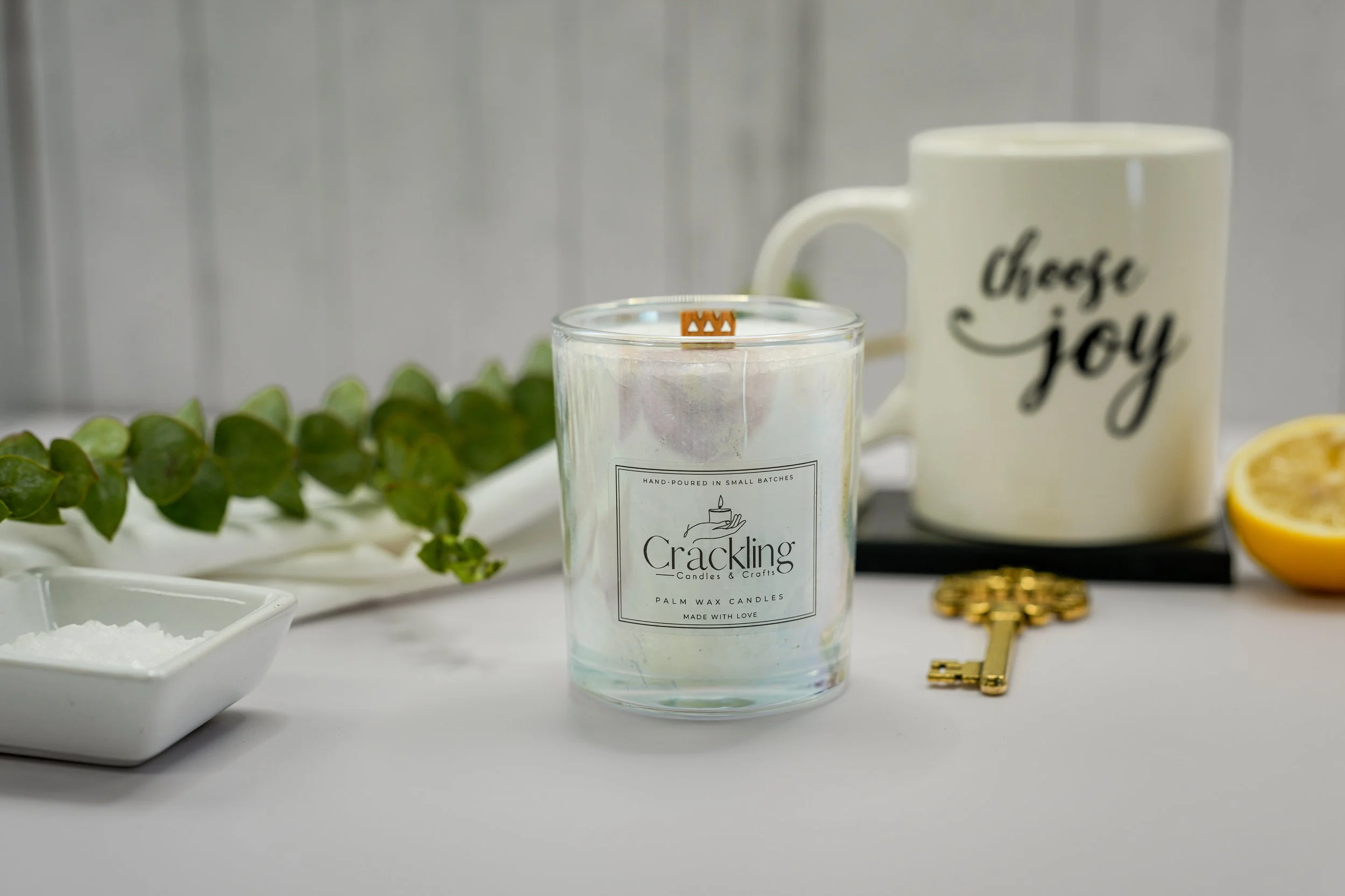 Blood Orange candle displayed beside a Choose Joy mug, antique brass key, and fresh green leaves in a bright neutral setting.