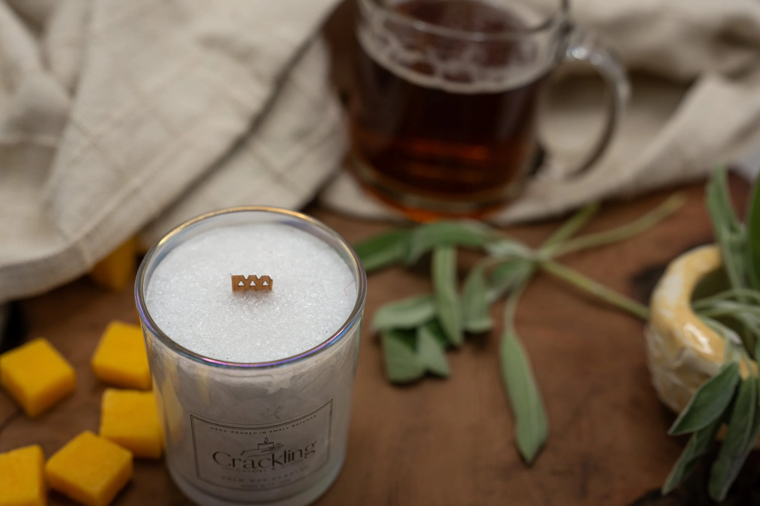 Angled overhead view of a palm wax candle showing its crystalline wax and wooden wick, with green leaves and a glass mug nearby.