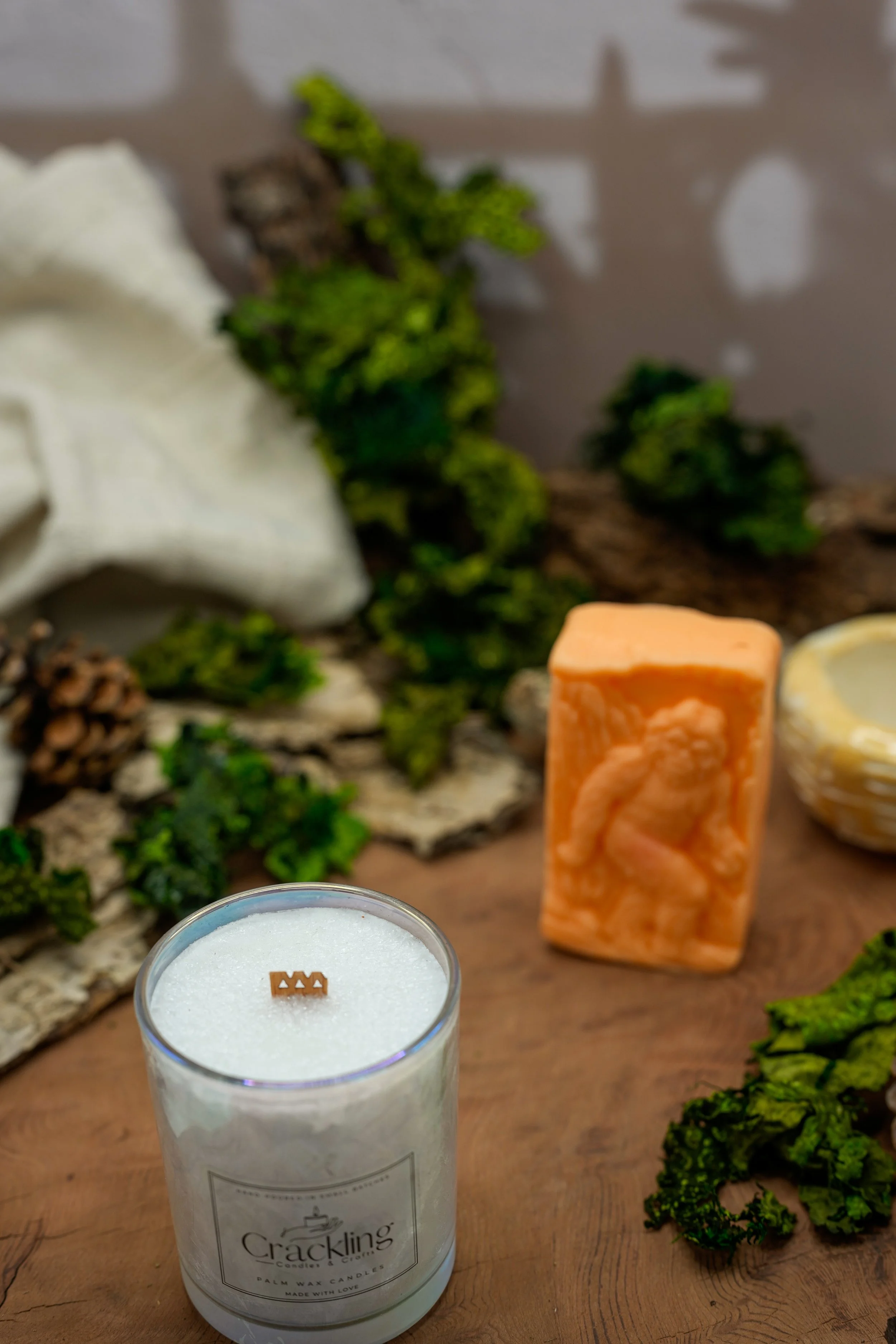 Cardamom & Teakwood candle photographed with rustic greenery, pinecones, cream linen, and warm natural accents.