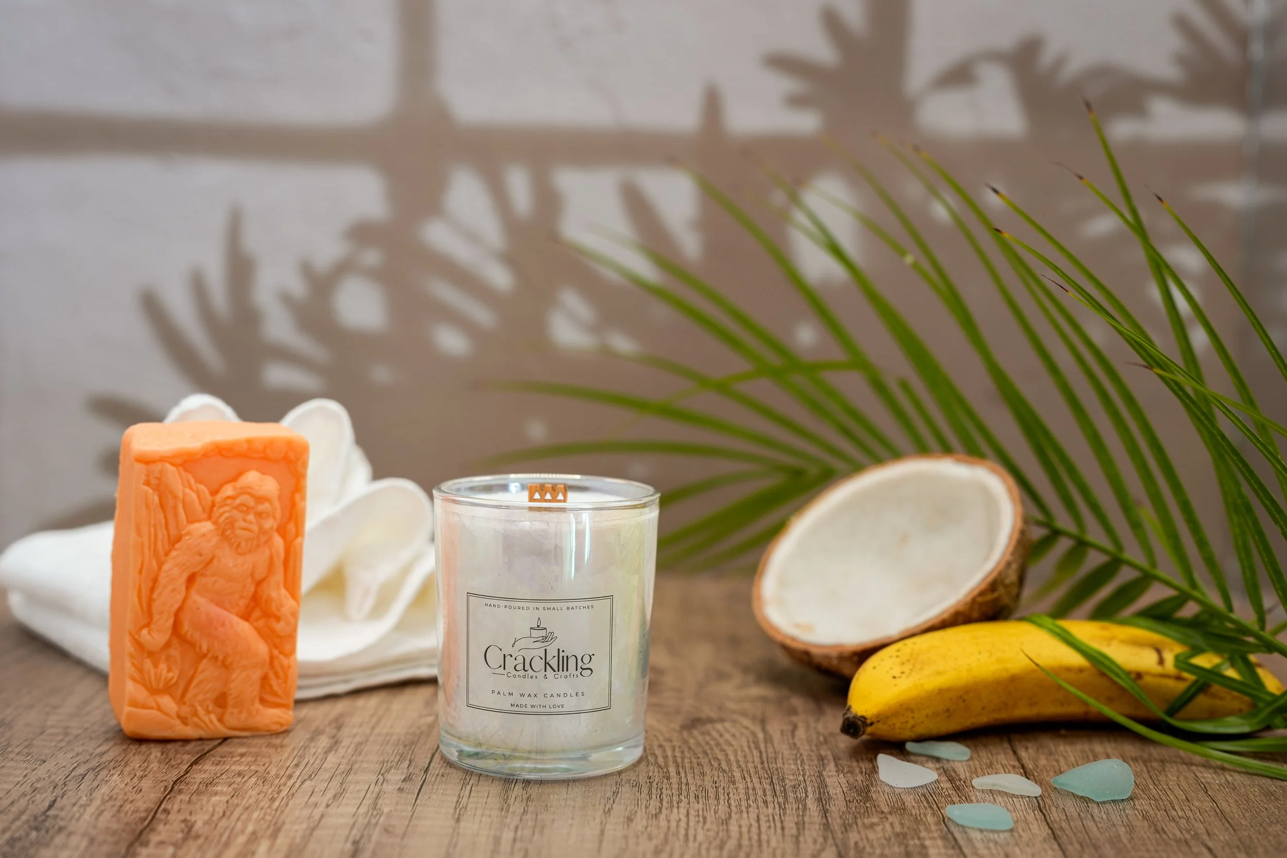 6 oz Butt Naked palm wax candle in a glass jar beside coconut, banana, and tropical fruit with palm leaves in the background.