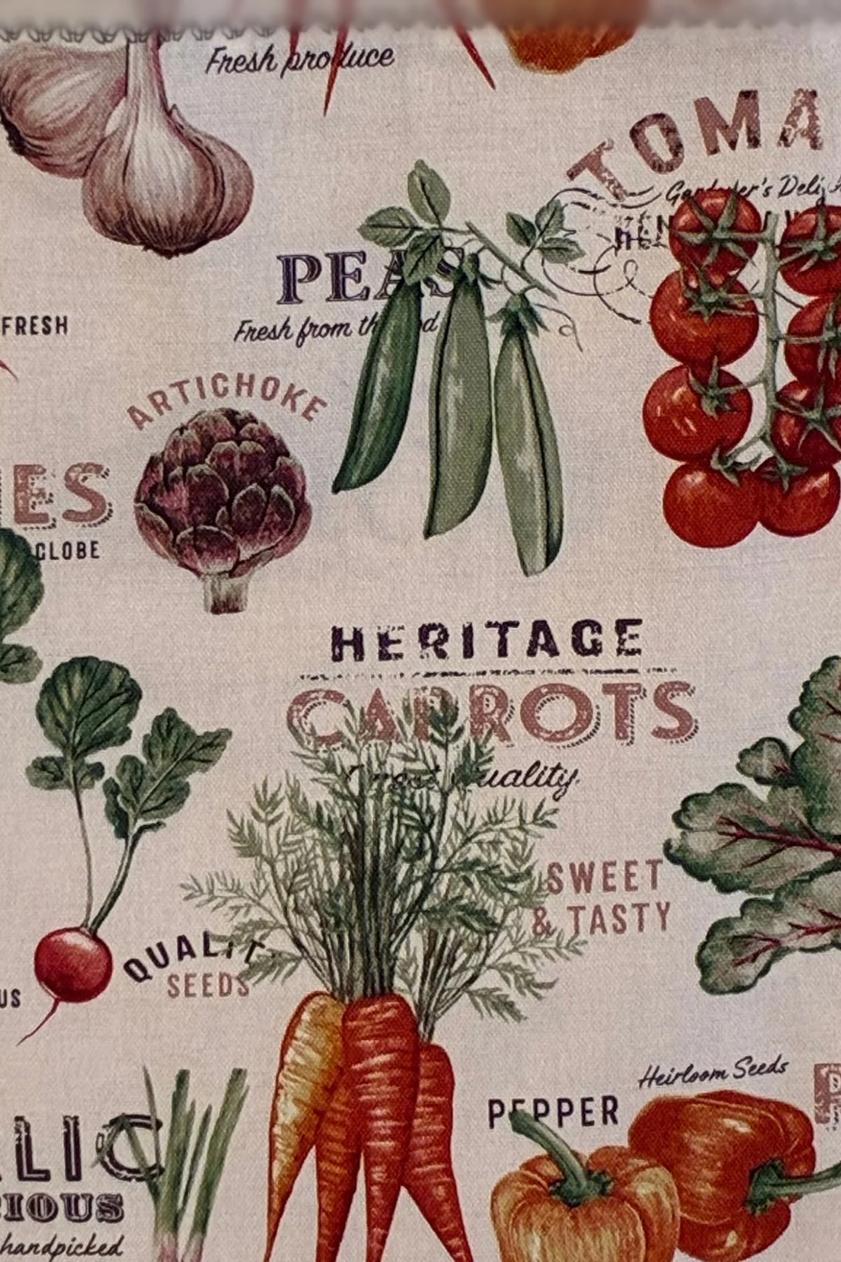 Close-up of cotton fabric featuring illustrated carrots, peas, tomatoes, and garden-inspired lettering on a light background.