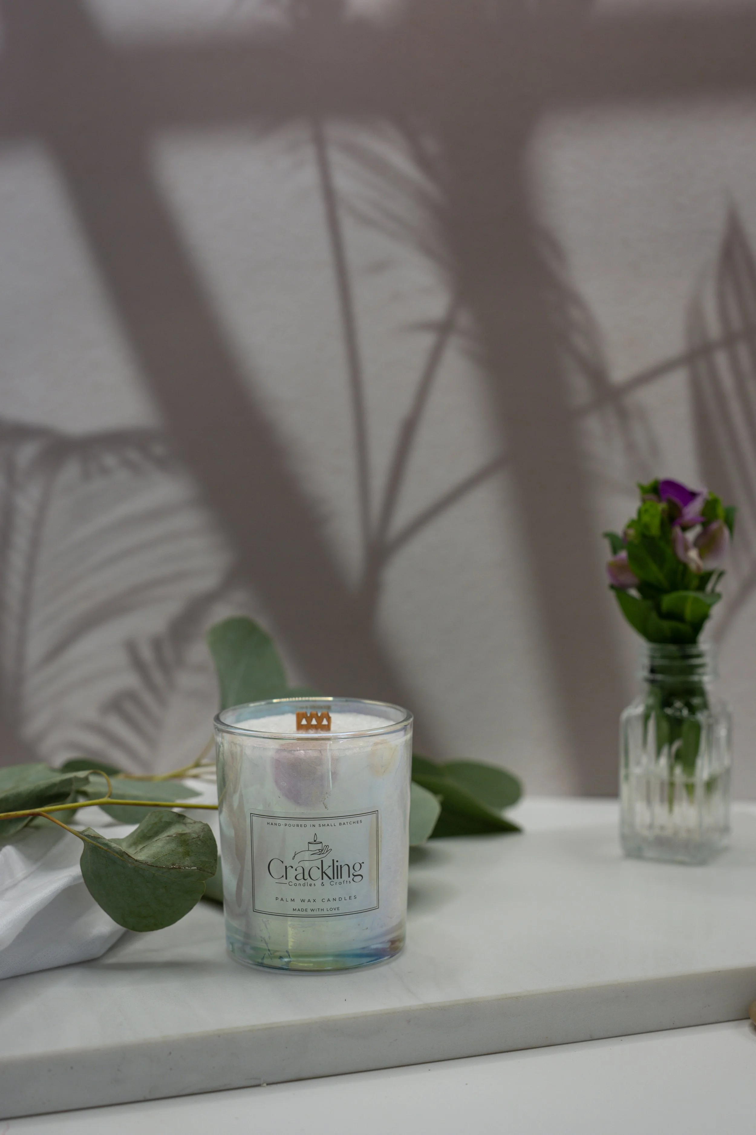 Sweet Pea candle displayed with eucalyptus leaves and a small vase of purple flowers against a warm neutral backdrop.