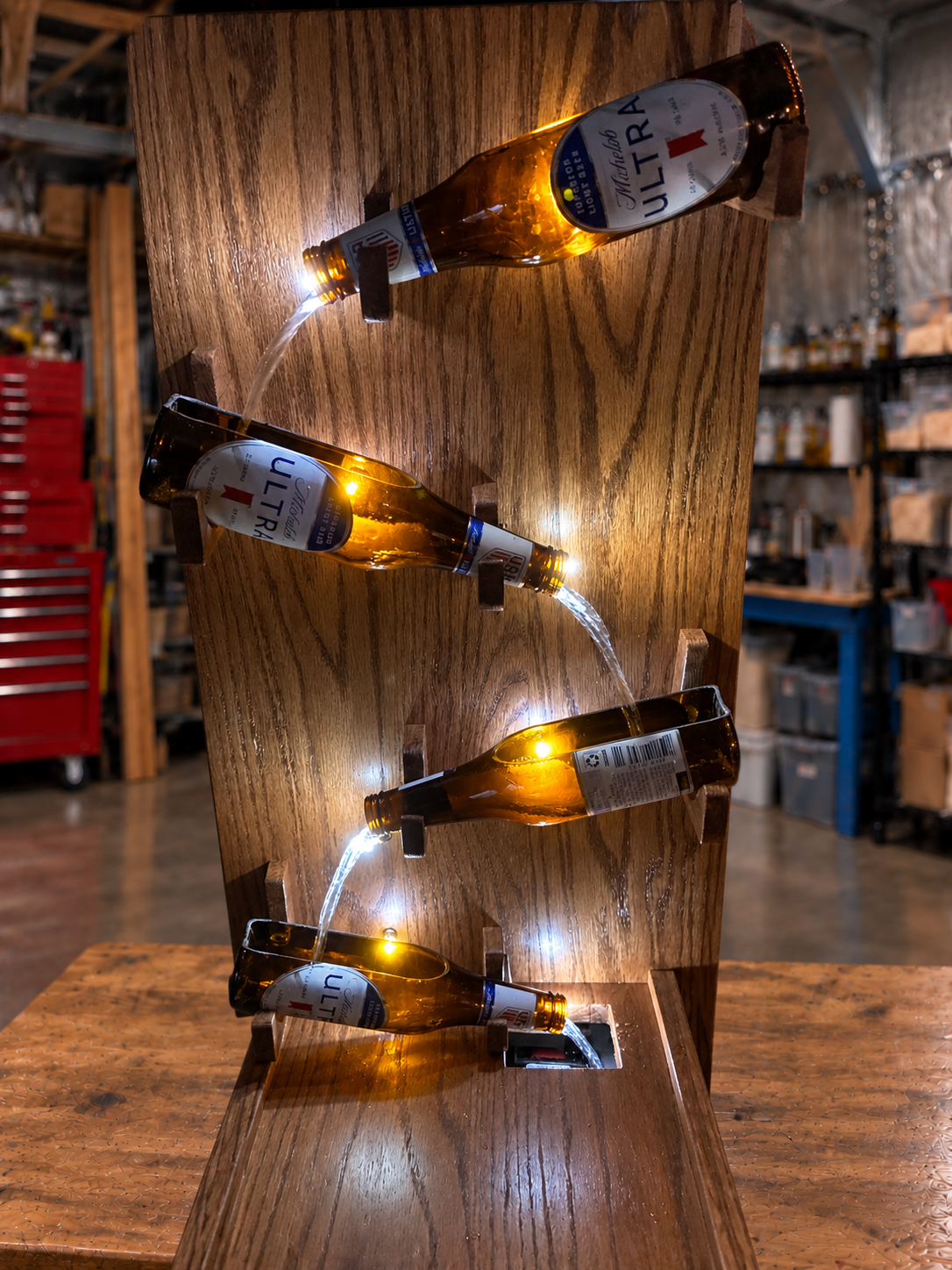 Close-up of cut Michelob Ultra bottles cascading water down a red oak fountain, showing illuminated bottle openings, wooden supports, and the water return opening.