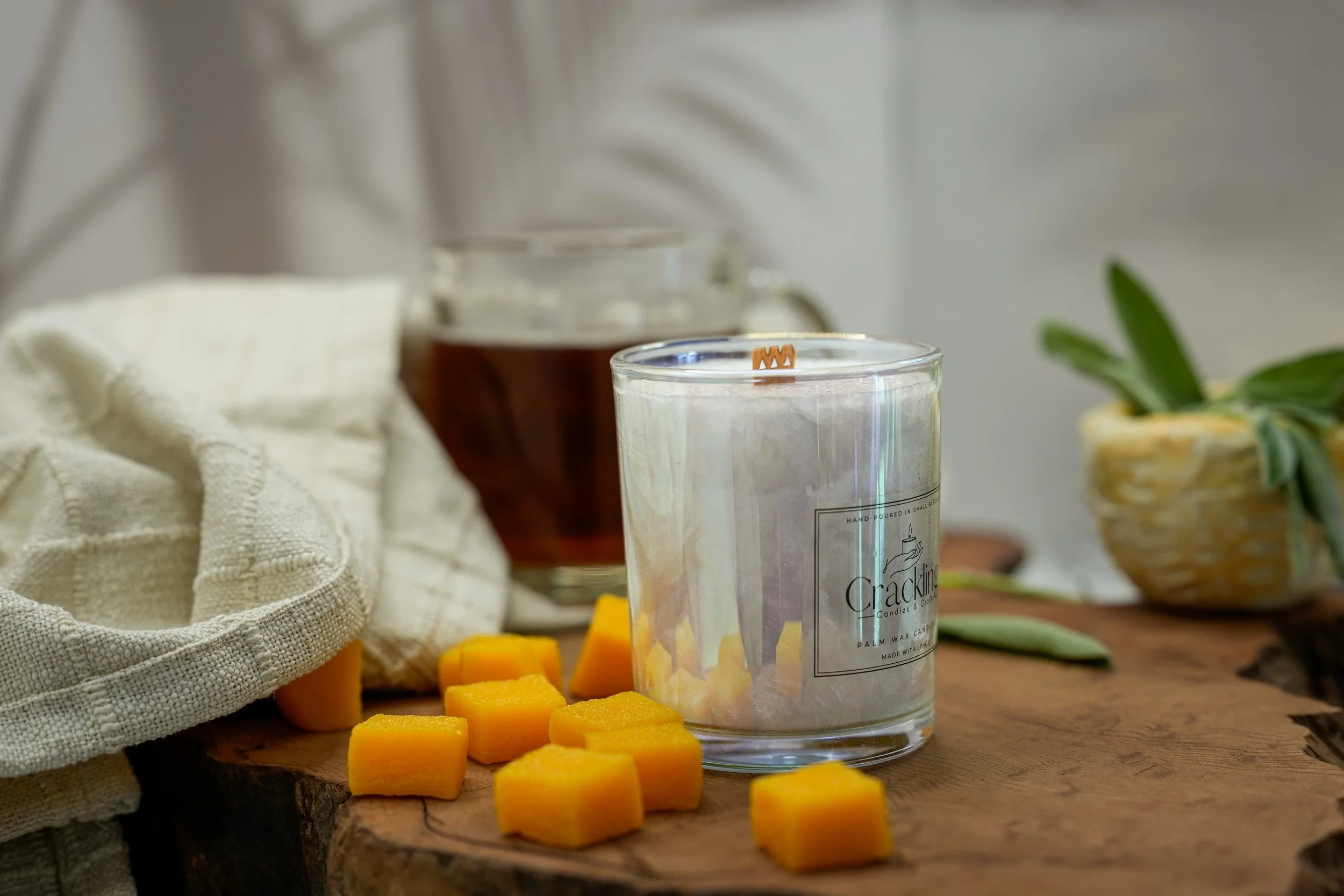 Palm wax candle in a clear glass jar beside mango cubes, sage leaves, and a glass mug on a wooden board.