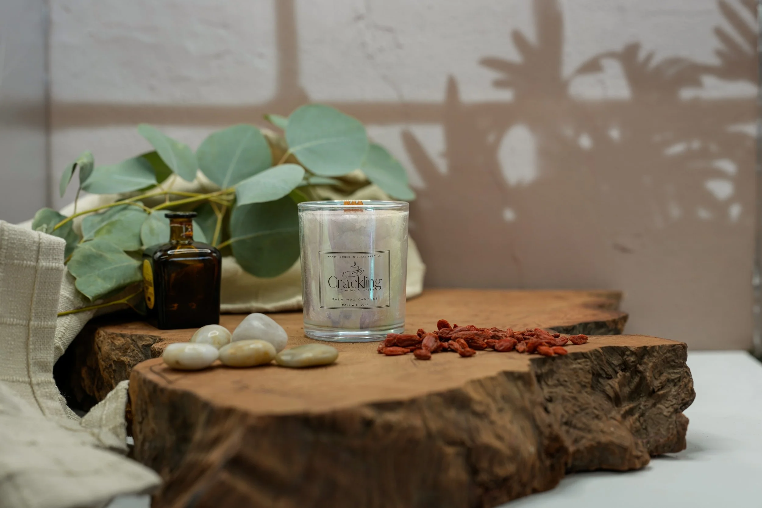 6 oz Tobacco & Oud palm wax candle with a wooden wick, styled on a natural wood slab with eucalyptus, smooth stones, and a small amber bottle