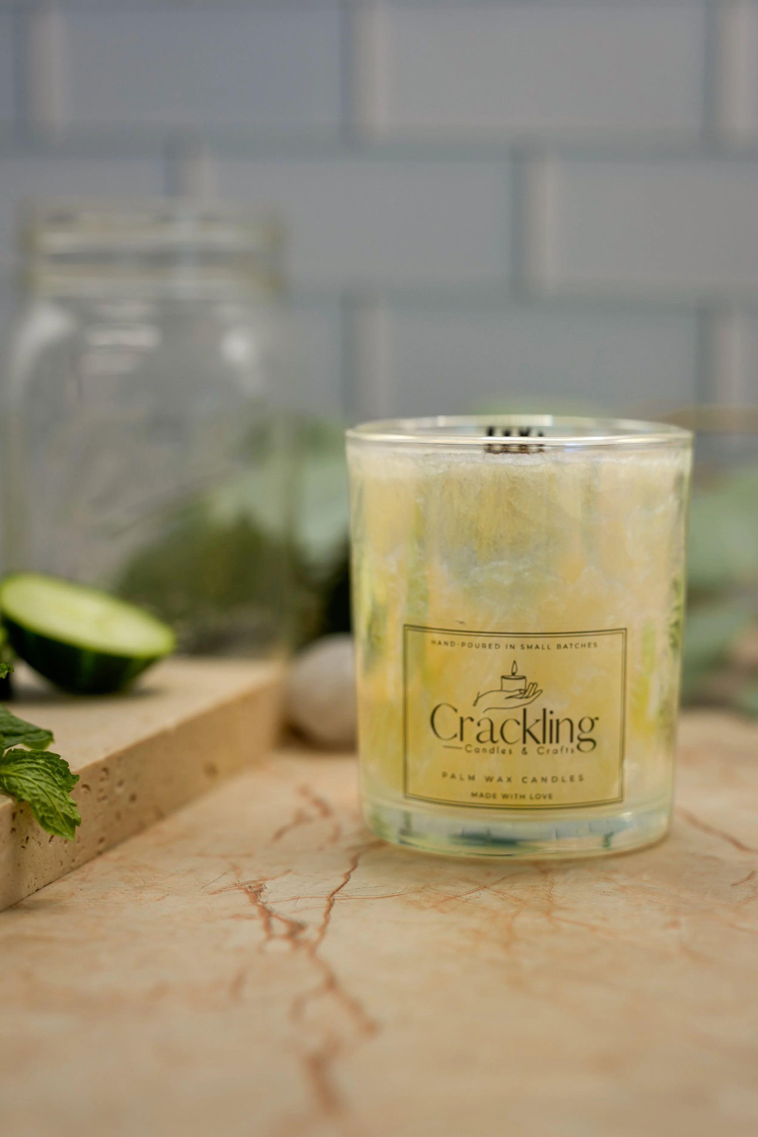 Close-up of the Garden Mint & Cucumber candle in a glass jar on a beige marble-patterned surface, styled with cucumber slices and green leaves.