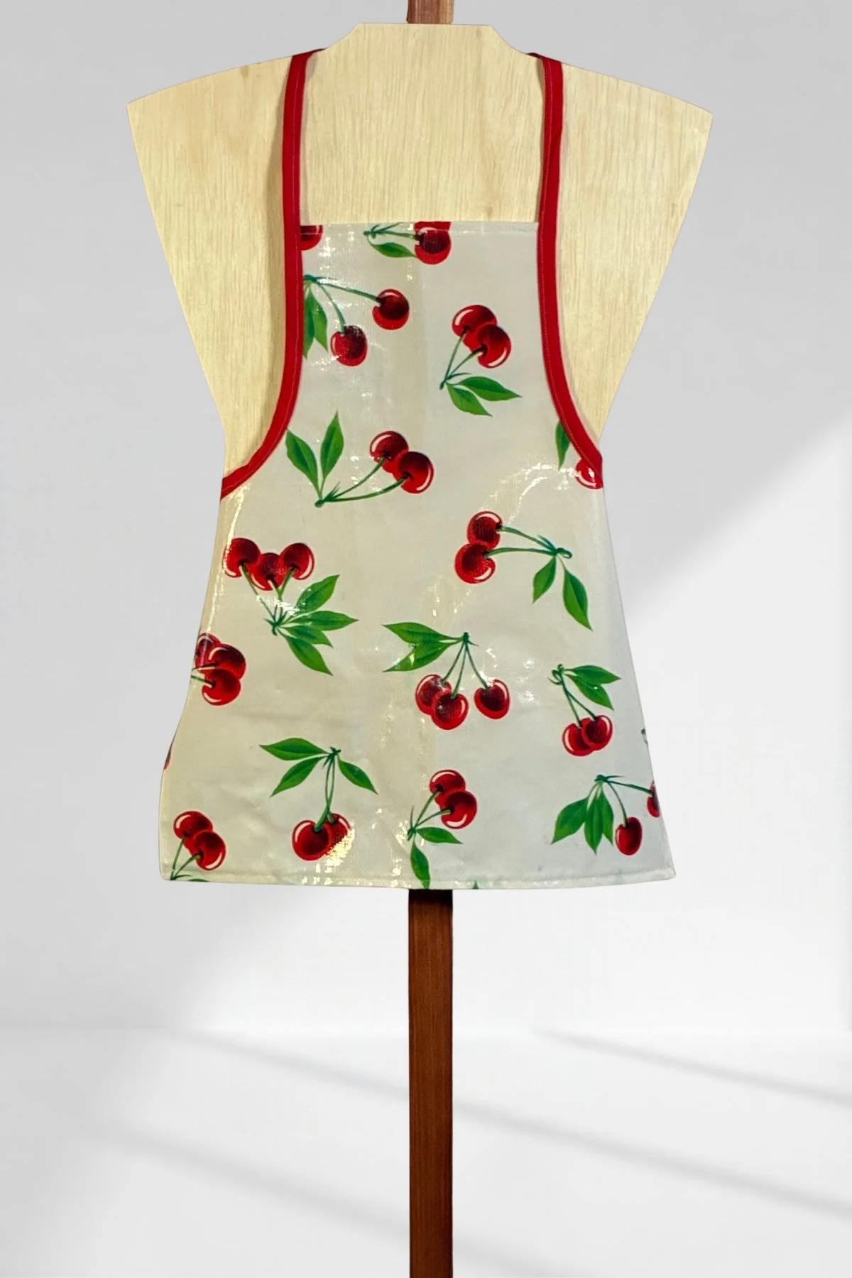 Handmade children’s oilcloth craft apron with red cherries, green leaves, and red trim, displayed on a wooden stand.