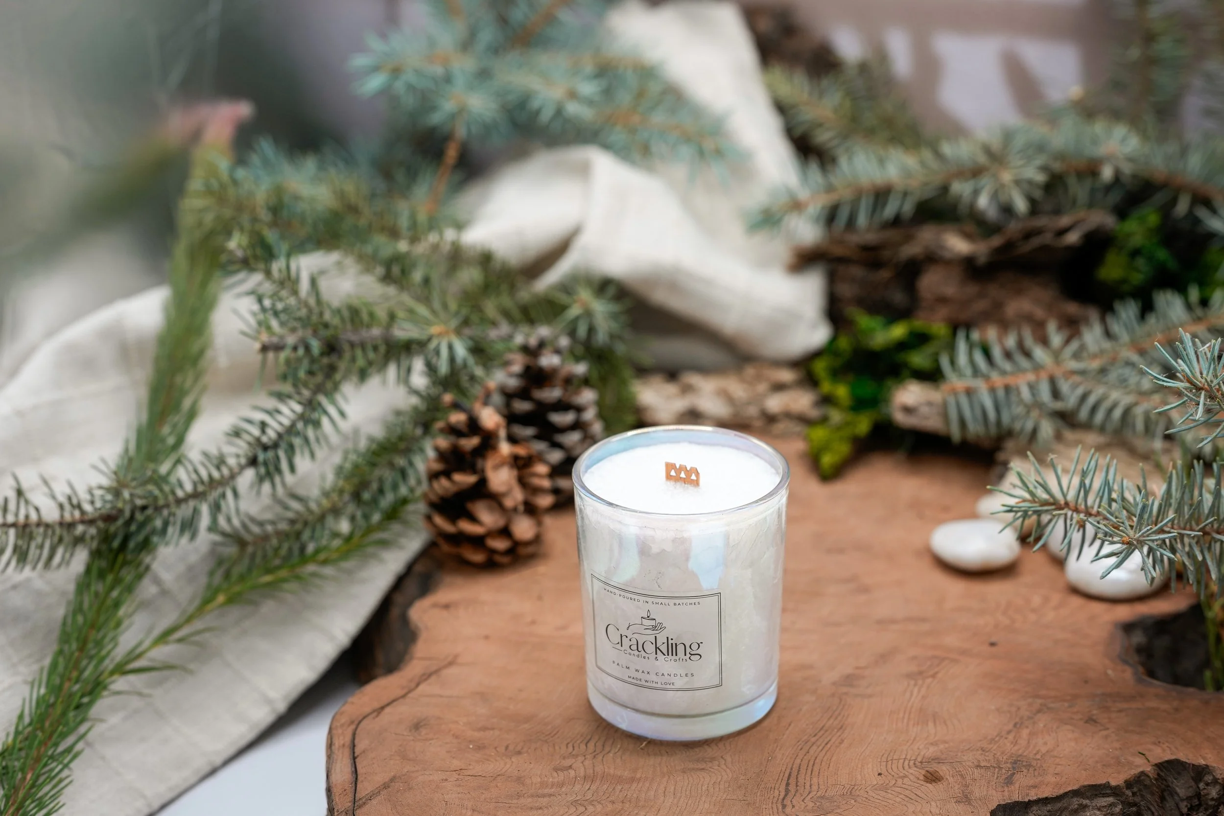 Angled view of the Forest Pine candle showing white wax and a wooden wick, styled on natural wood with evergreen branches, a pinecone, and cream fabric.
