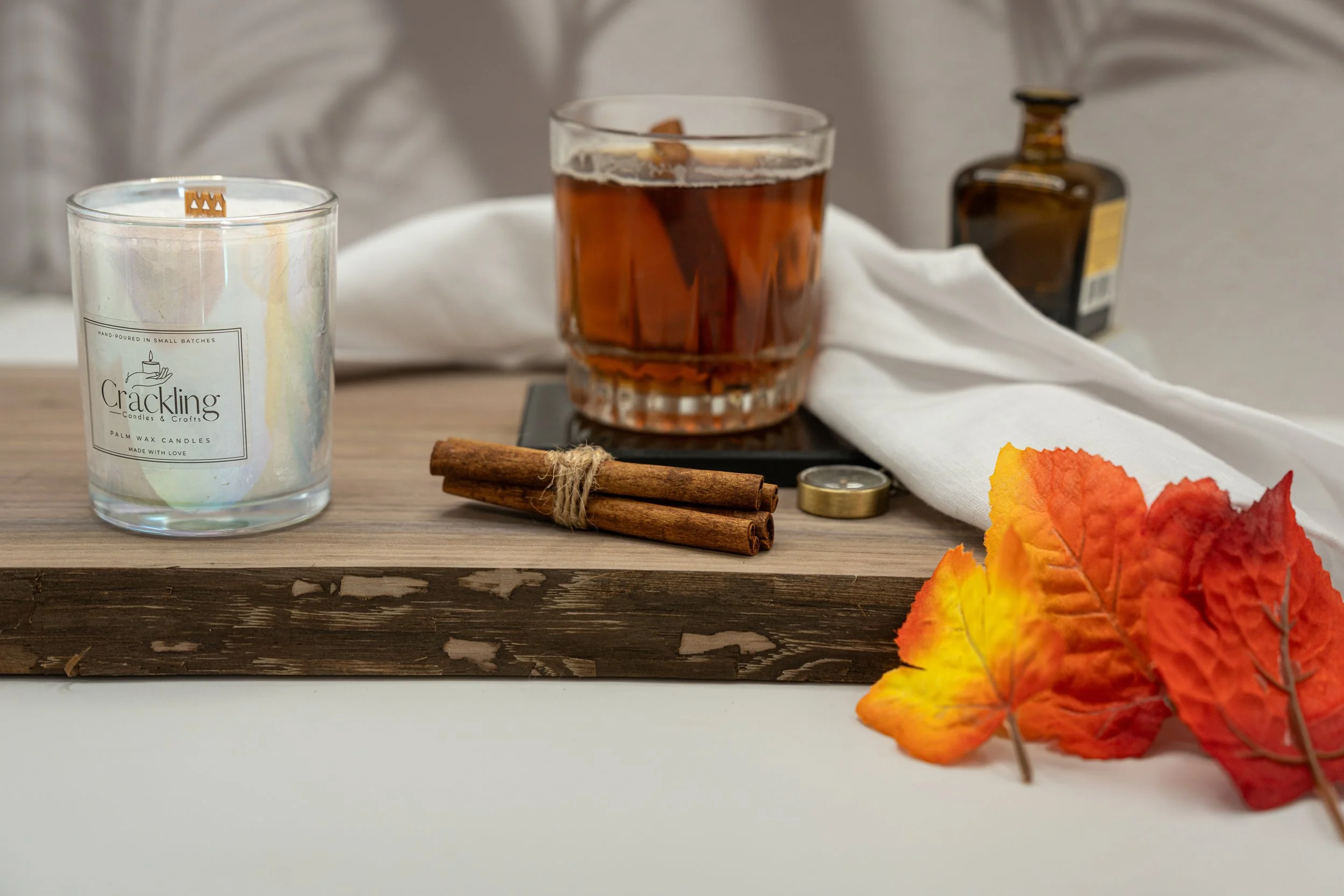 Maple Whiskey Toddy candle partially visible beside an amber drink, cinnamon sticks, and autumn leaves on a wooden board.