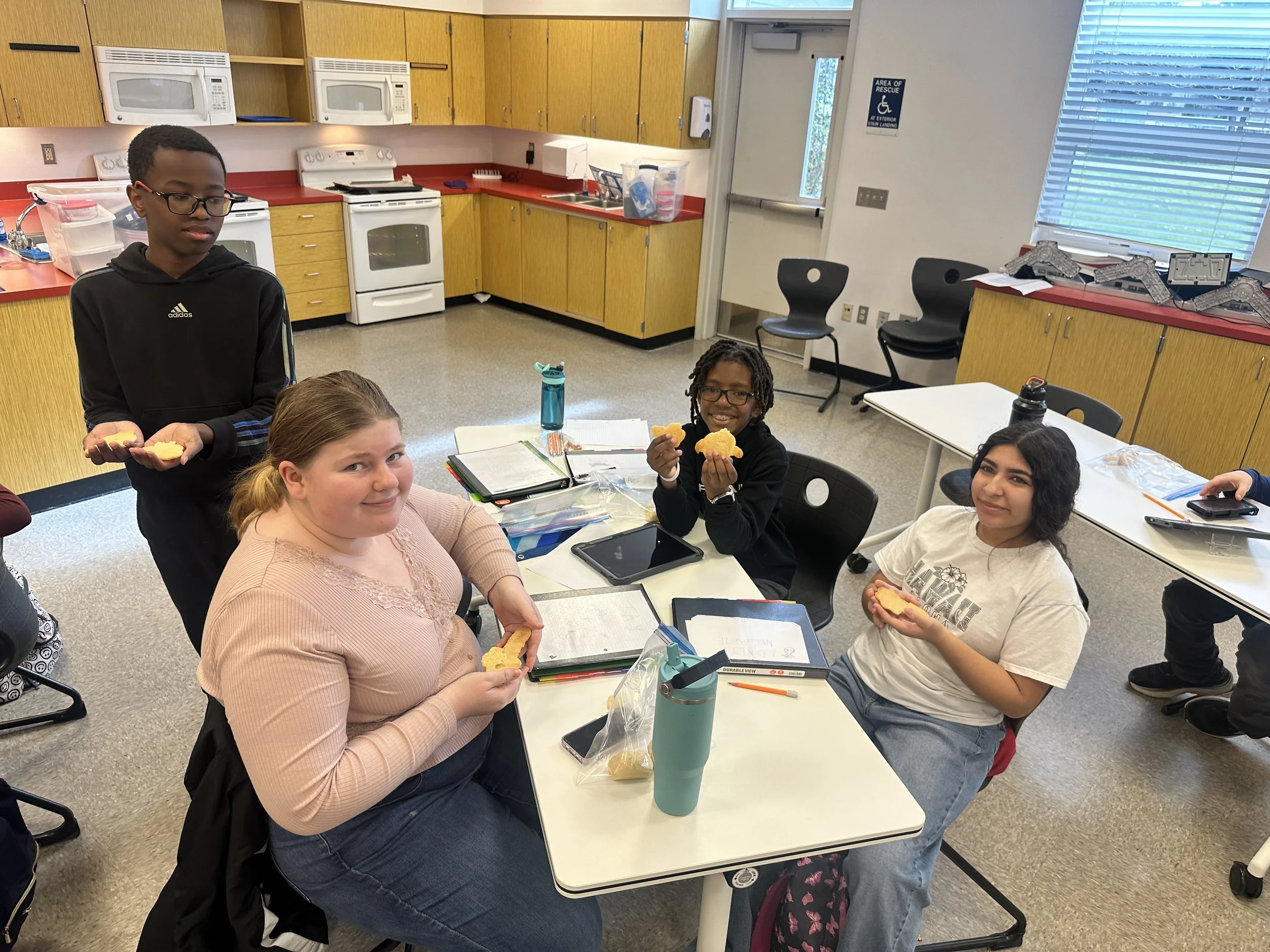 A group of diverse children sitting at a table in a kitchen or classroom, enjoying snacks and smiling at the camera. The room has wooden cabinets, white appliances, and large windows with blinds.